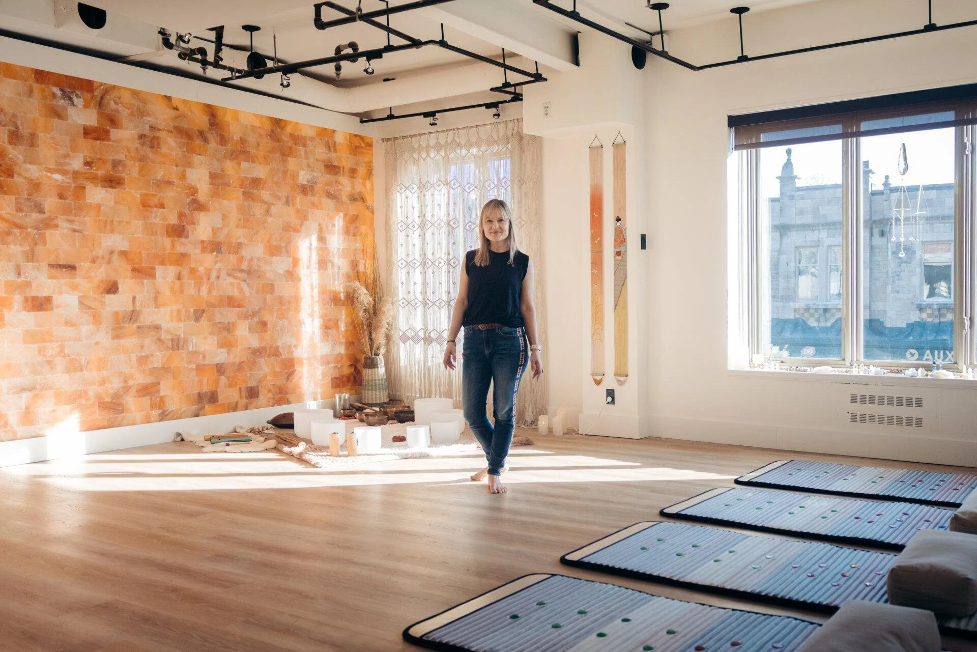 A woman standing barefoot in a bright, modern room with a wooden floor, orange brick accent wall, and large windows. The room is decorated with candles, a woven basket with dried plants, and various ritual items on the floor.