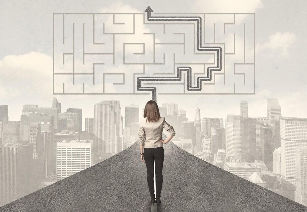 A woman stands on a city rooftop looking at a wall-sized maze and a black path leading through it to the top center, symbolizing finding a path through challenges.