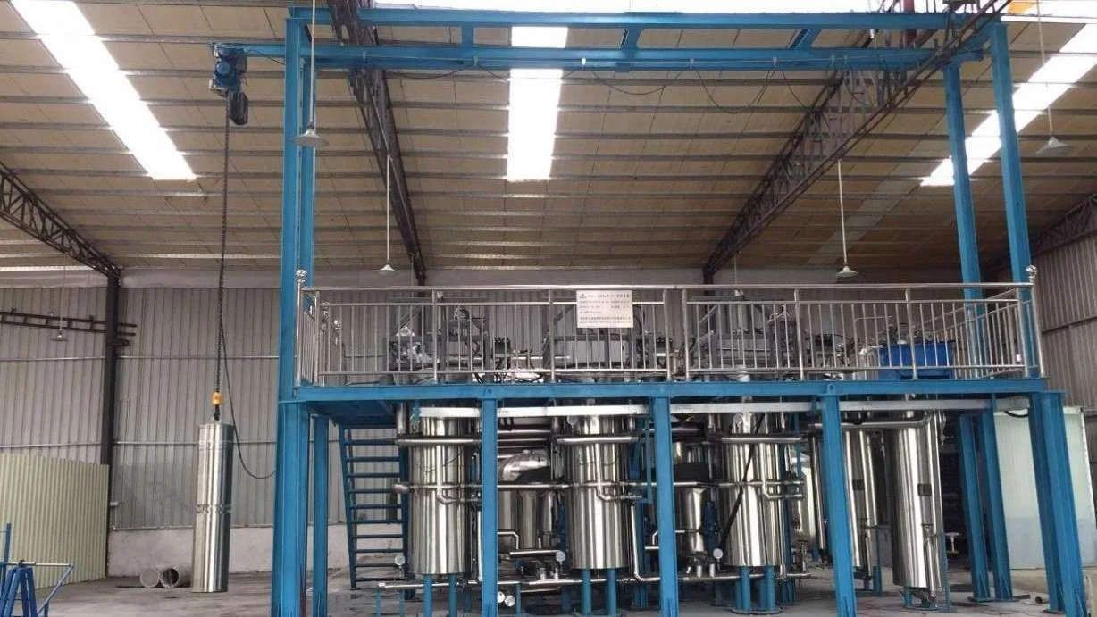 Industrial facility with a two-story structure, metallic tanks, and pipes, used for processing or manufacturing. The upper level has a safety railing and some equipment, while the lower level contains large cylindrical tanks connected by pipes.