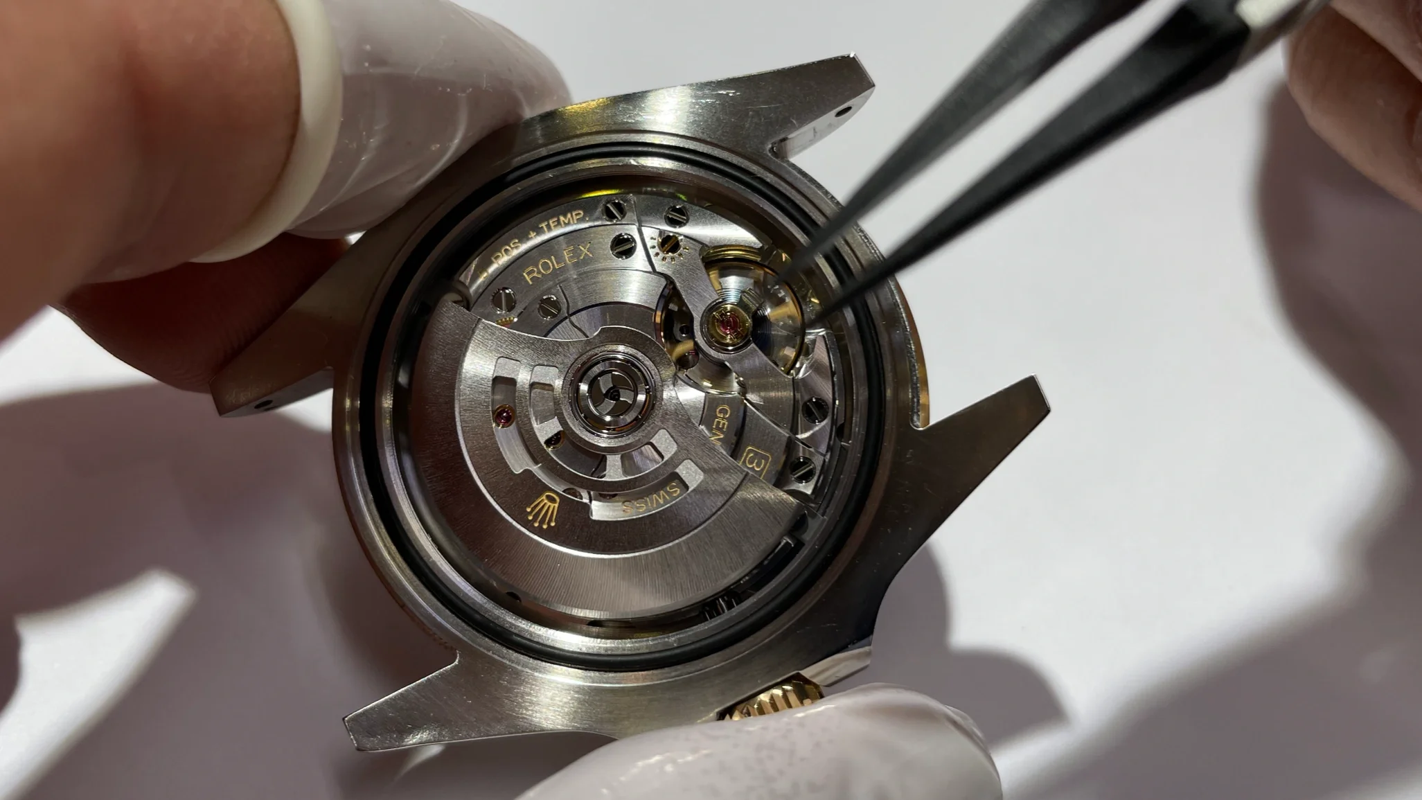 Close-up of a watchmaker's gloved hands working on the intricate mechanical movement of a Rolex watch, with screws and gears visible.