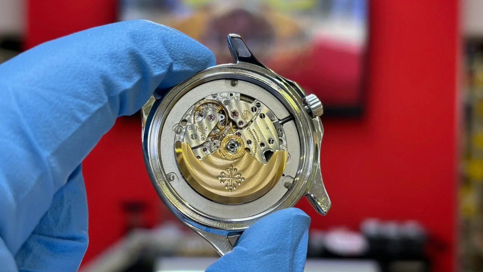 Close-up of a luxury watch movement with intricate gears and a gold rotor, held by a gloved hand, in front of a red background.