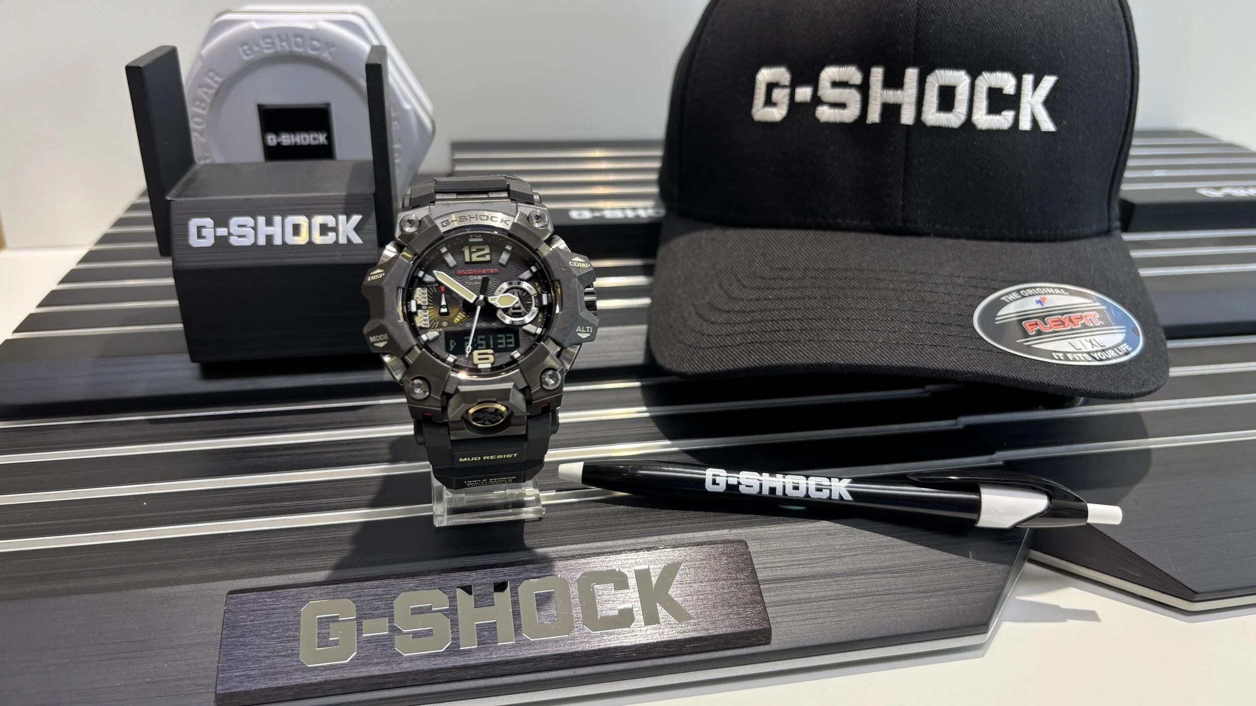 A G-Shock watch, a black cap with 'G-SHOCK' embroidered, a black G-Shock pen, a sticker with 'G-SHOCK', a display box, and a stand with a G-Shock logo.