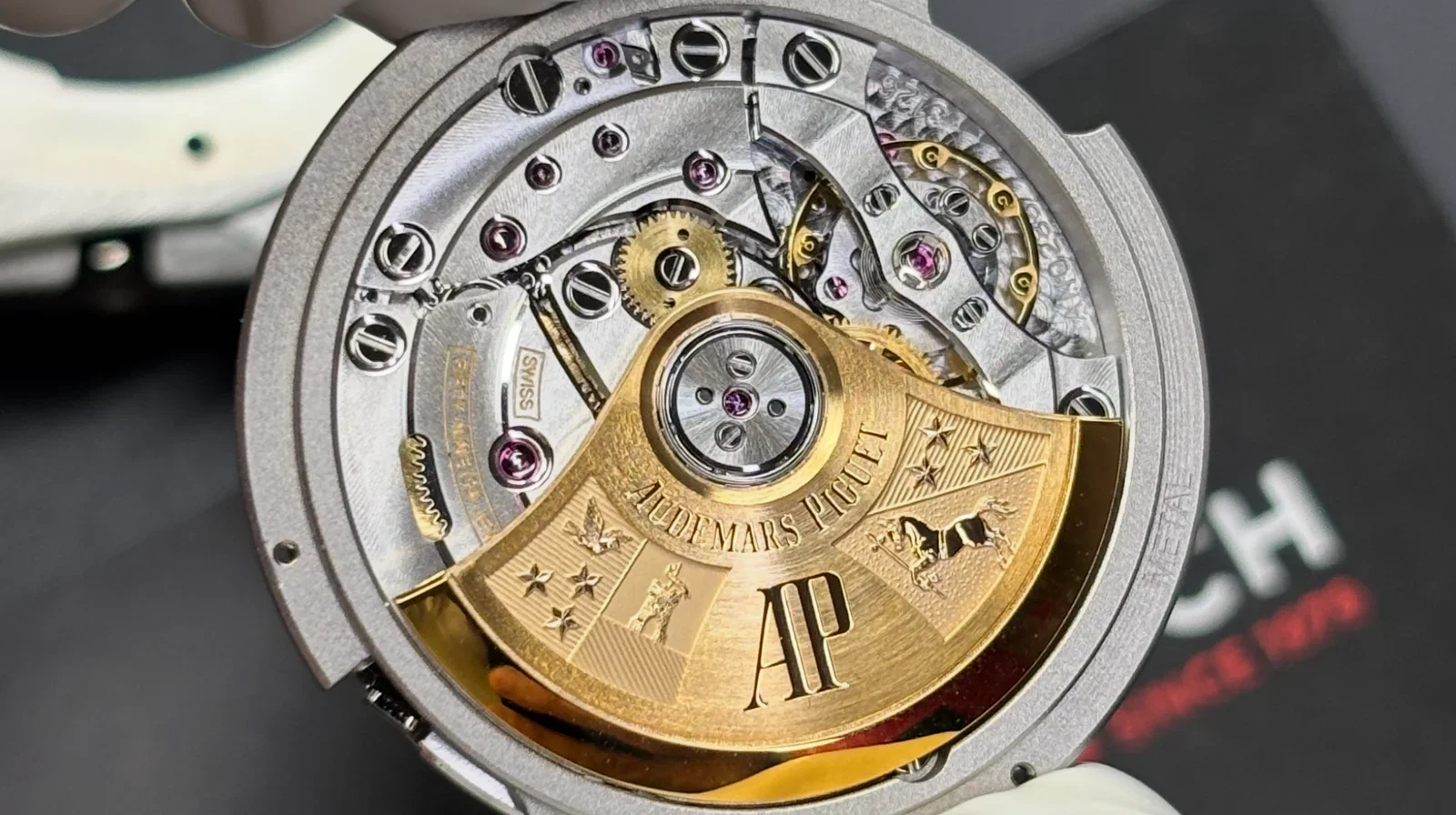 Close-up of a luxury watch movement with intricate gears and components, featuring a gold rotor engraved with the name Audemars Piguet and a large letter 'AP'.