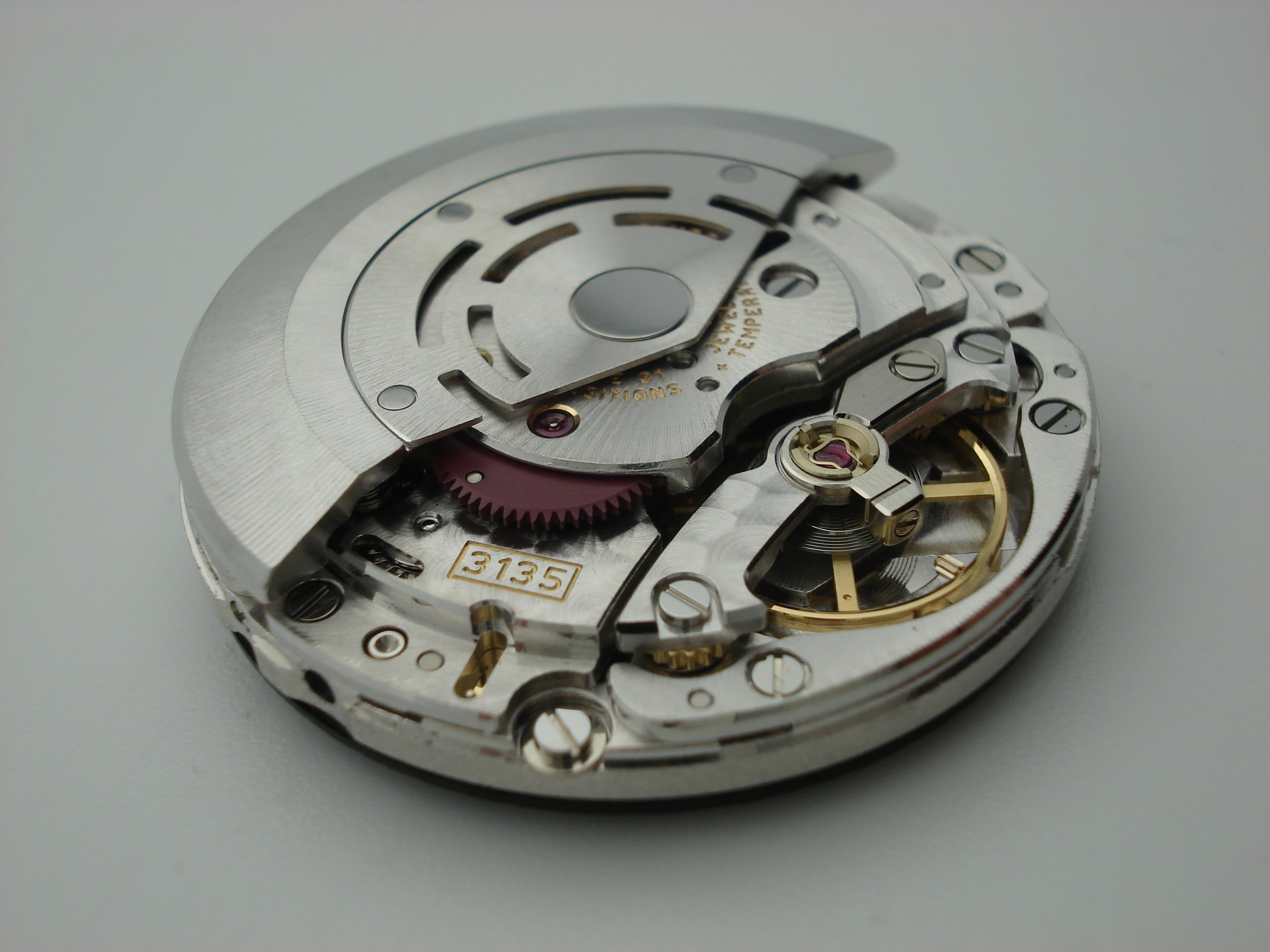 Close-up of a mechanical watch movement with various gears, screws, and components, including a pink gear and gold details.
