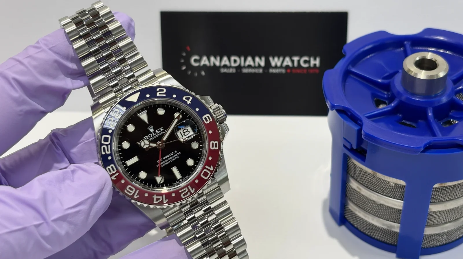 A person wearing a purple glove holding a Rolex GMT-Master II watch with a black dial and a two-tone red and blue bezel. In the background, there is a black card with the logo and name 'Canadian Watch' and a blue watchmaker's holder or polishing mach