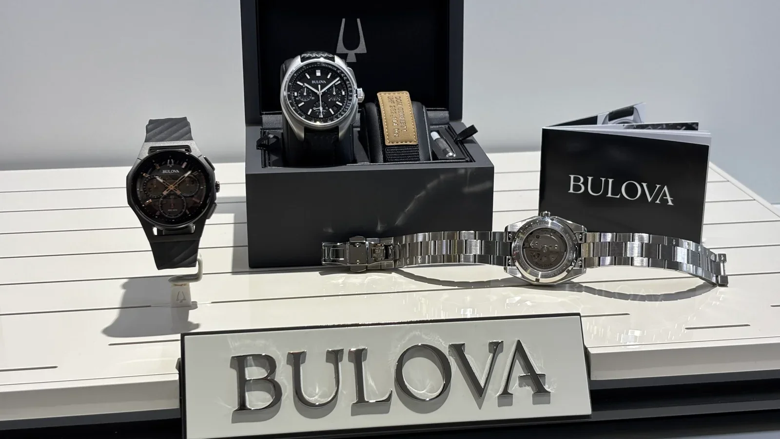 Display of Bulova watches and accessories on a white surface, including two wristwatches, a box with additional watch components, and a Bulova brochure.