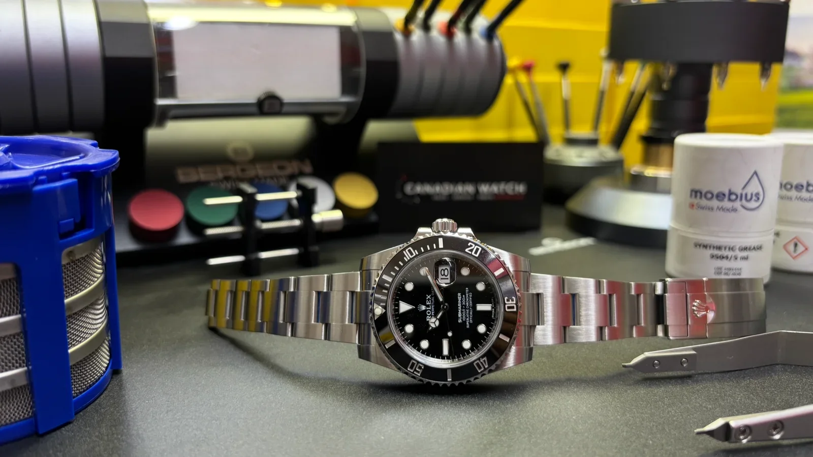 A Rolex Submariner watch with a black dial and rotating bezel on a workbench surrounded by watch tools, lubricant, and watch parts.