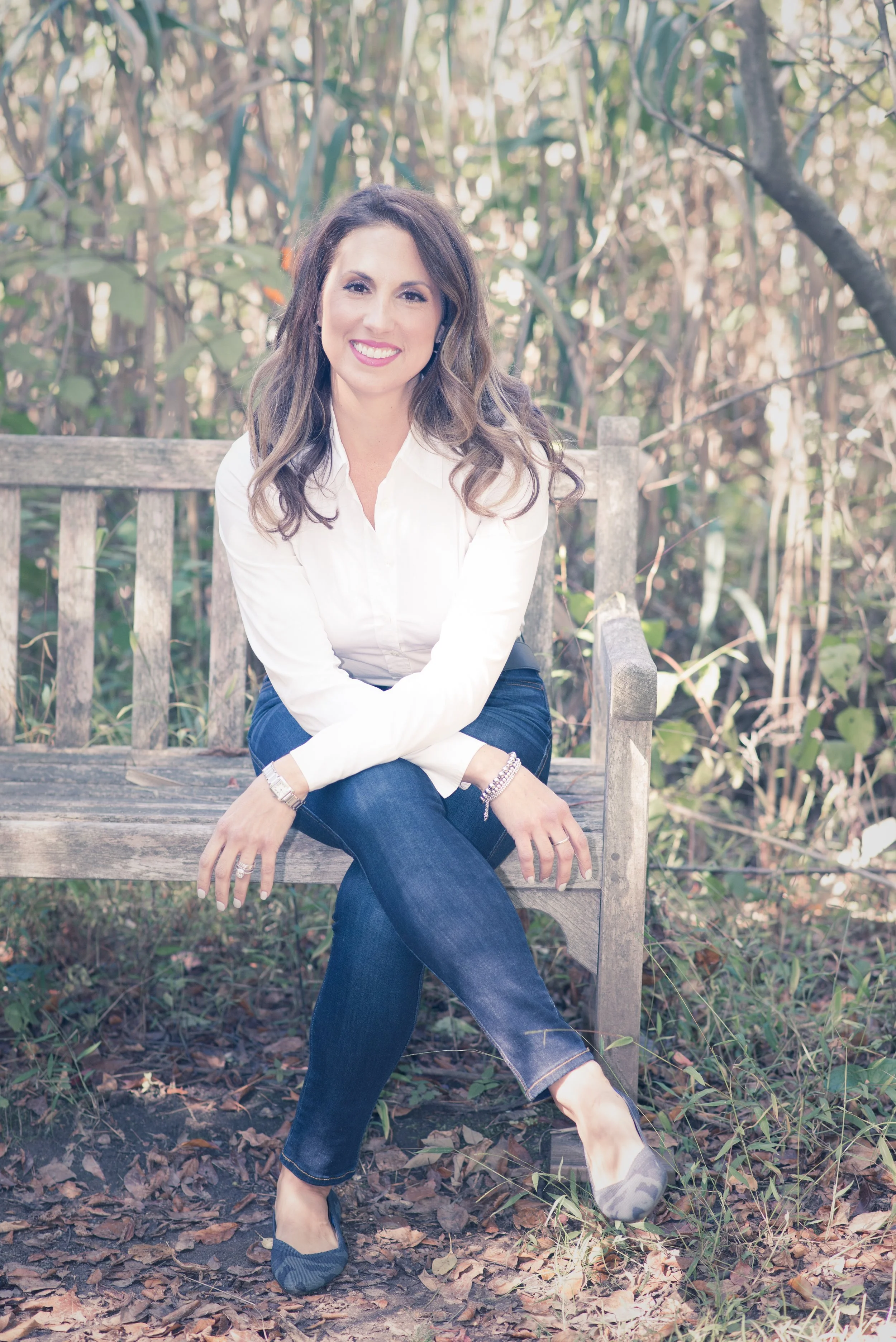 A woman with long brown curly hair and light skin is sitting on a wooden bench outdoors in a wooded area. She is wearing a white blouse, blue jeans, and slip-on shoes. She is smiling, with her hands resting on her lap and one leg crossed over the other. The background includes trees and leaves on the ground.