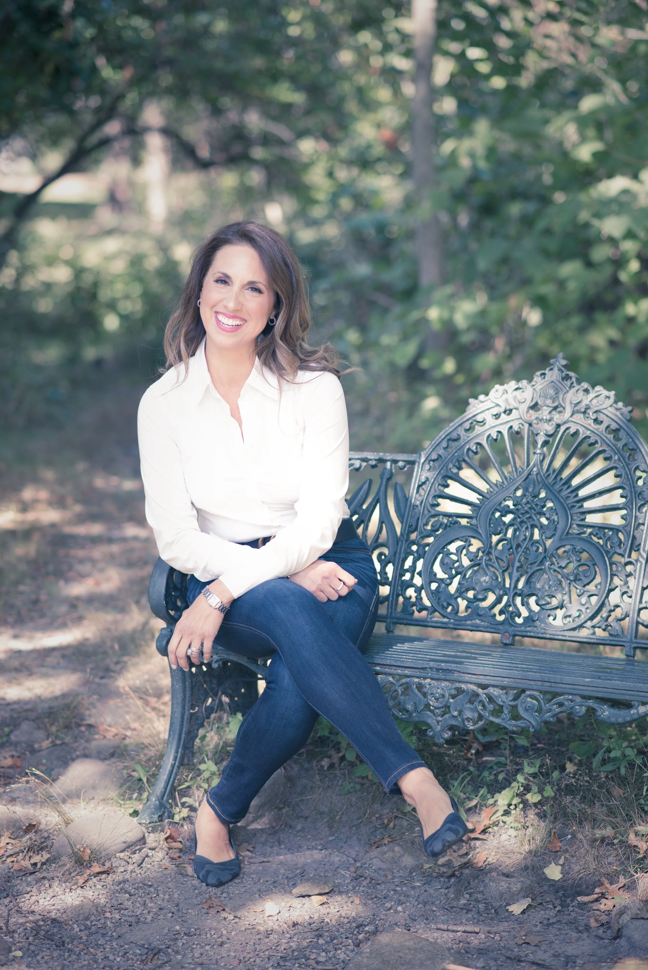 A woman with dark wavy hair sitting on a decorative black metal park bench outdoors, smiling, wearing a white blouse, dark jeans, and navy flats, surrounded by trees and natural scenery.