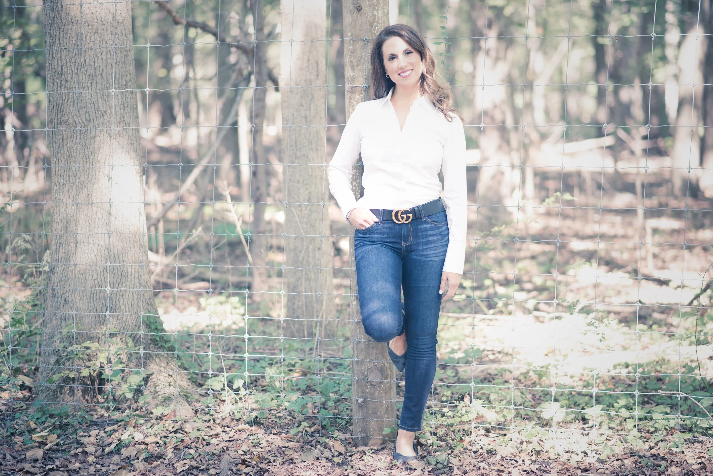 A woman with long brown hair, wearing a white blouse, blue jeans with a Gucci belt, and black shoes, stands in front of a wire fence in a wooded area, smiling at the camera.