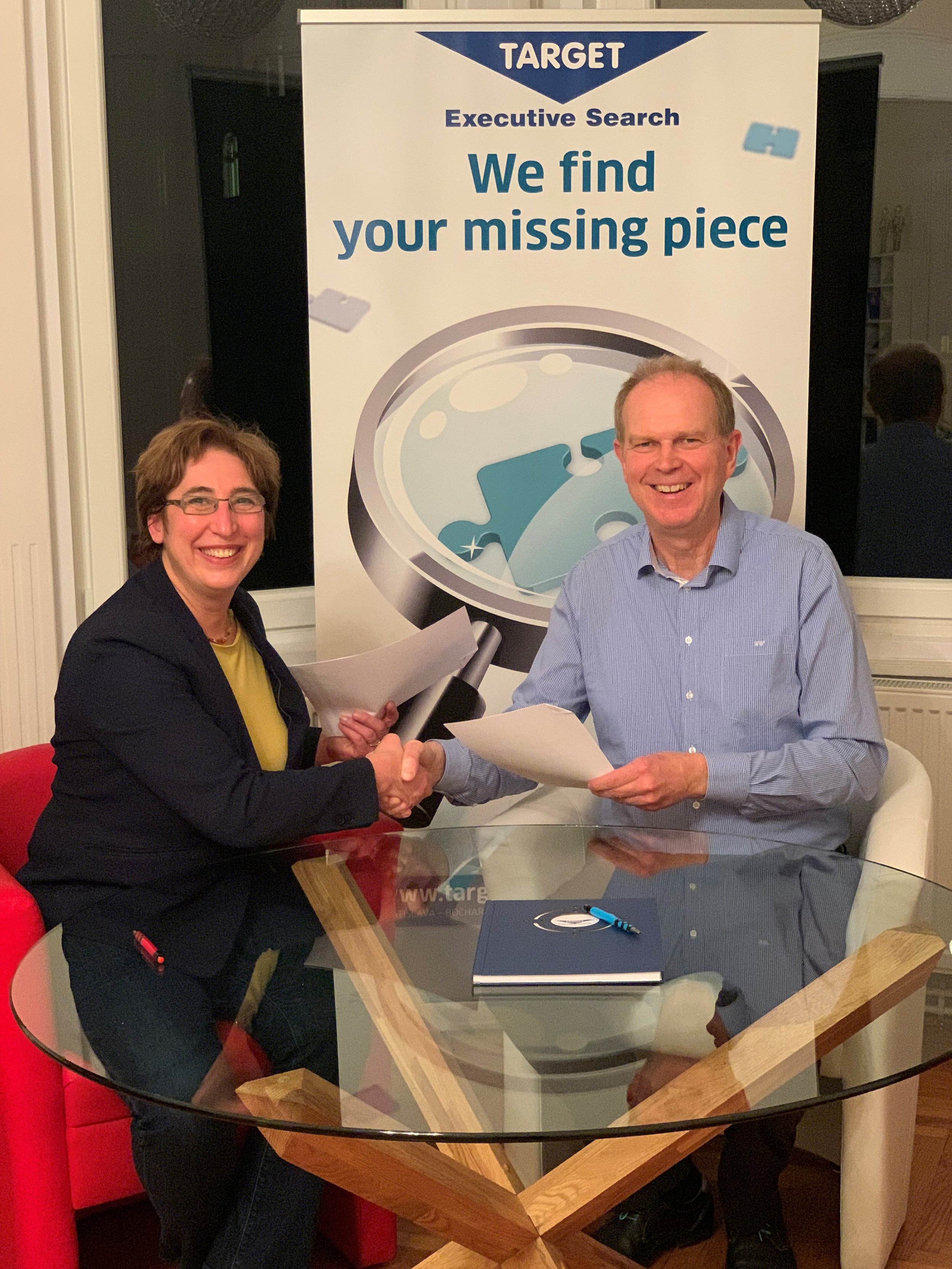 A woman and a man sitting at a glass-top table, shaking hands. Behind them is a large sign with the Target logo and the words 'Executive Search' and 'We find your missing piece.' The woman is holding a rolled-up paper, and there are papers and a navy-blue notebook on the table.