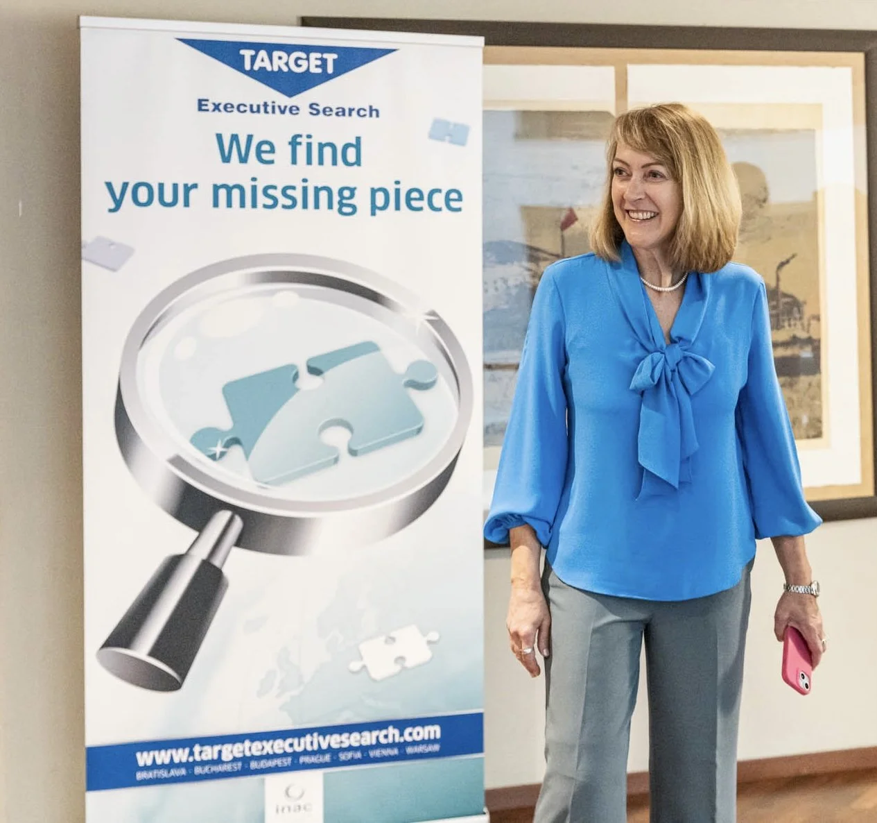 A woman in a blue blouse standing next to a Target-branded banner that reads, 'We find your missing piece,' with a magnifying glass focusing on puzzle pieces, in an office setting.