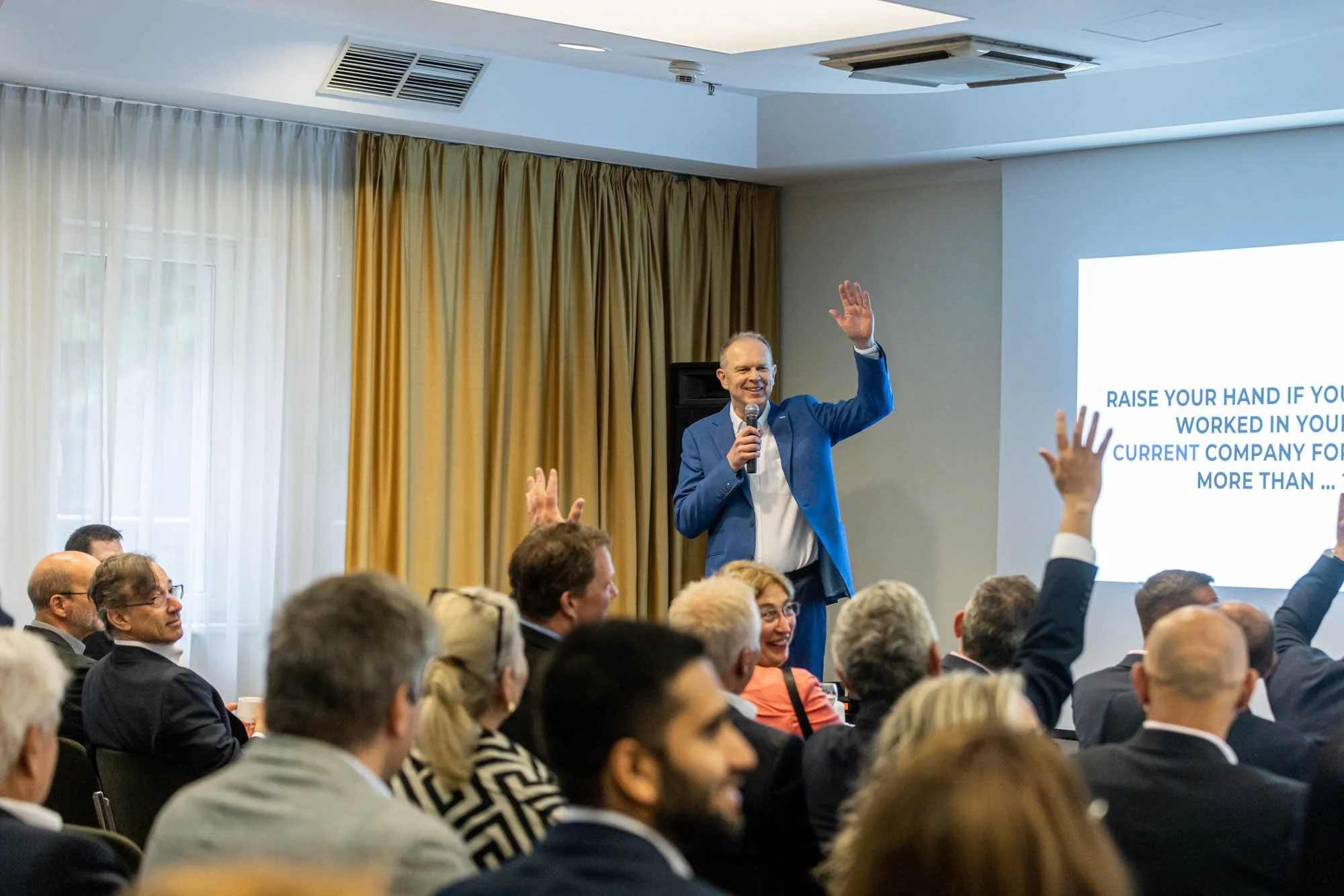 A man in a blue suit standing and speaking into a microphone during a conference or seminar, with audience members raising their hands.
