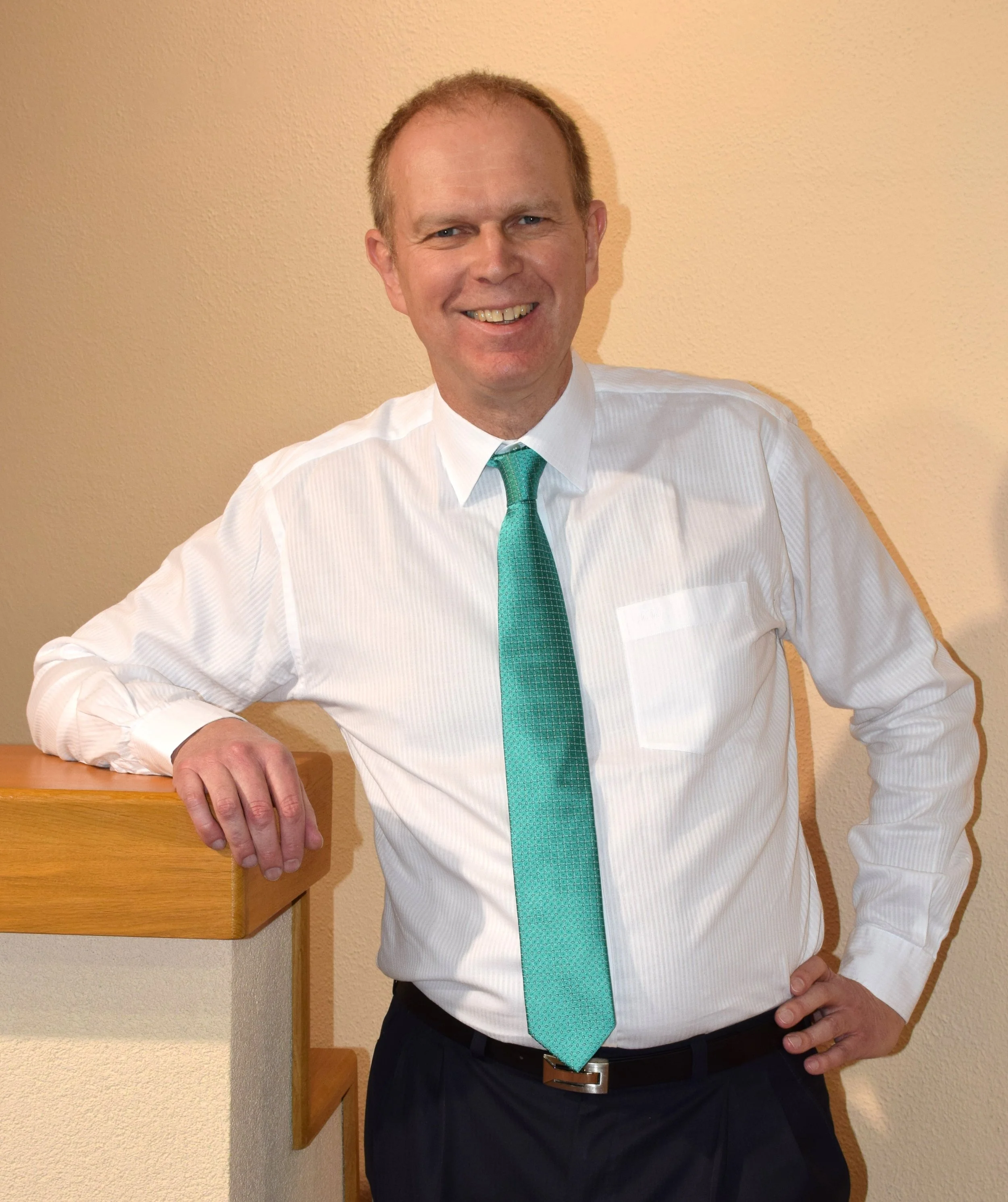 Klemens Wersonig, man in a white dress shirt, turquoise tie, and dark pants stands in front of a beige wall, smiling and resting his left hand on a wooden surface.