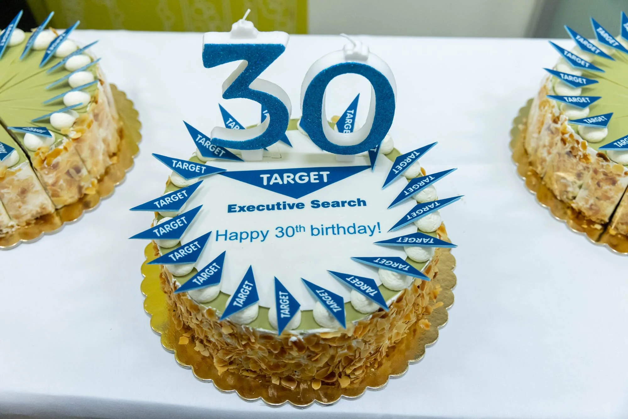 Cake decorated with blue 'target' flags, white frosting, and caramel-colored flakes. Contains blue number '30' candles, with the message 'Happy 30th birthday!' and 'Executive Search' written on the cake.