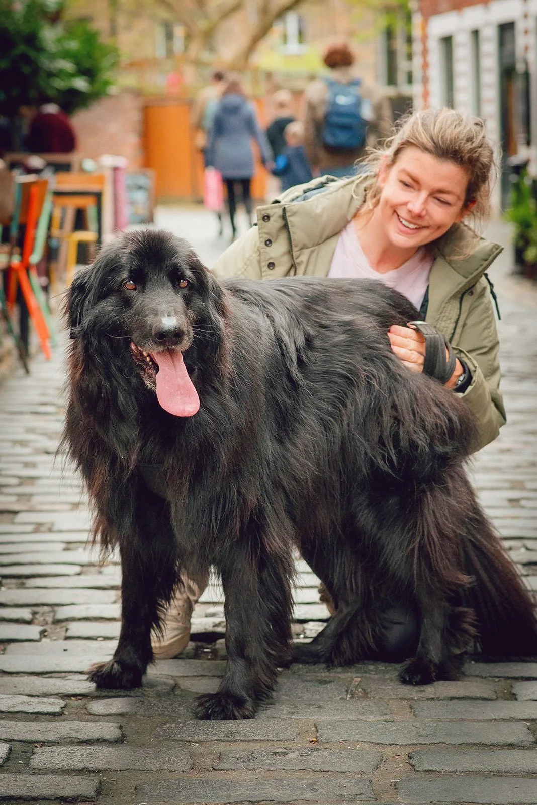 A female dog trainer with dark blonde hair, wearing a green jacket on a cobblestone street smiling at a German shepherd/bernese mix dog with a shiny coat and tongue hanging out, they are on Ashton Lane, in Glasgow's west end. Both look happy.