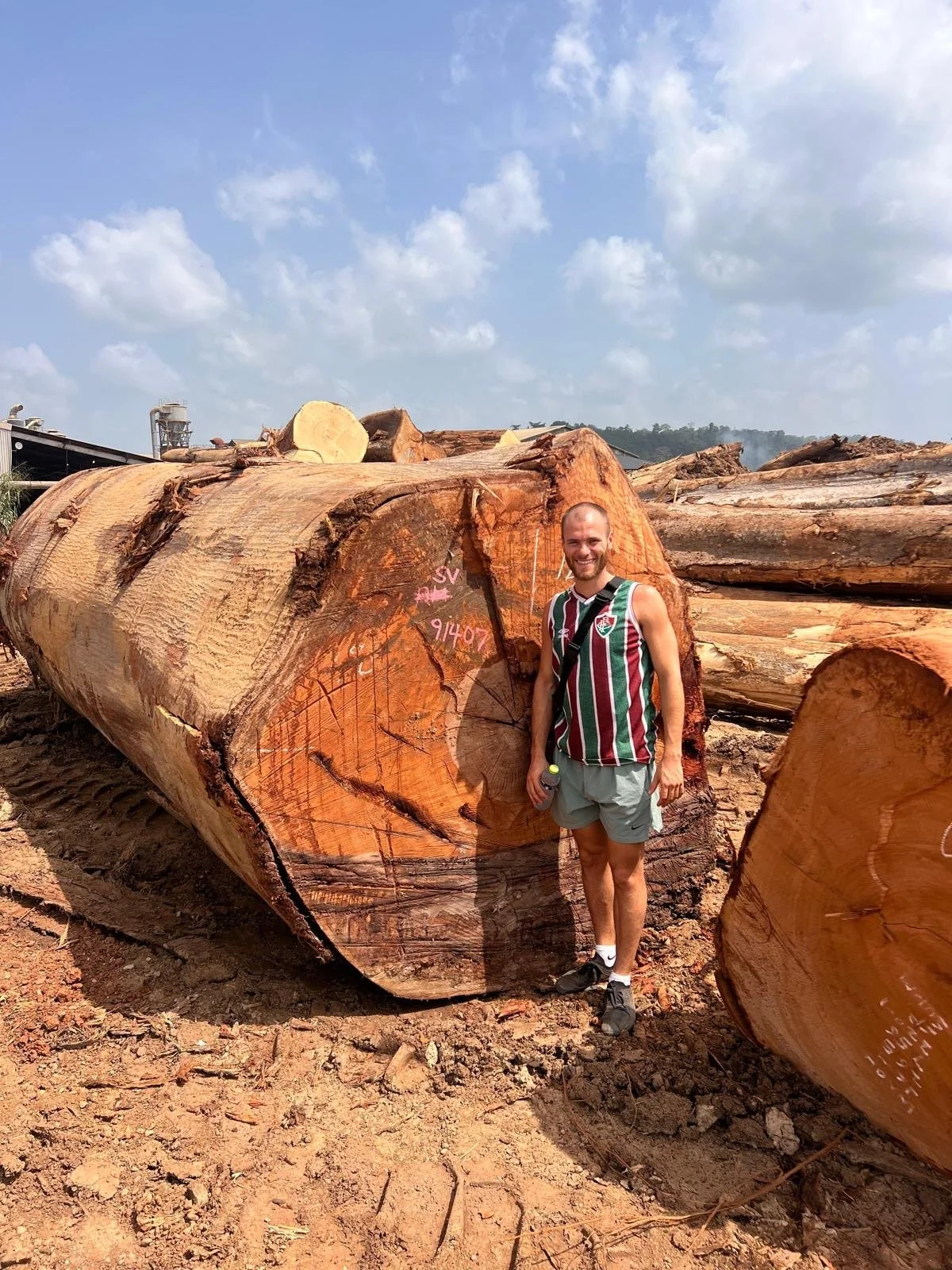 A man with a beard, wearing a striped sports jersey and shorts, standing next to a large cut log in a lumber yard or forest. He is holding a water bottle and smiling at the camera.