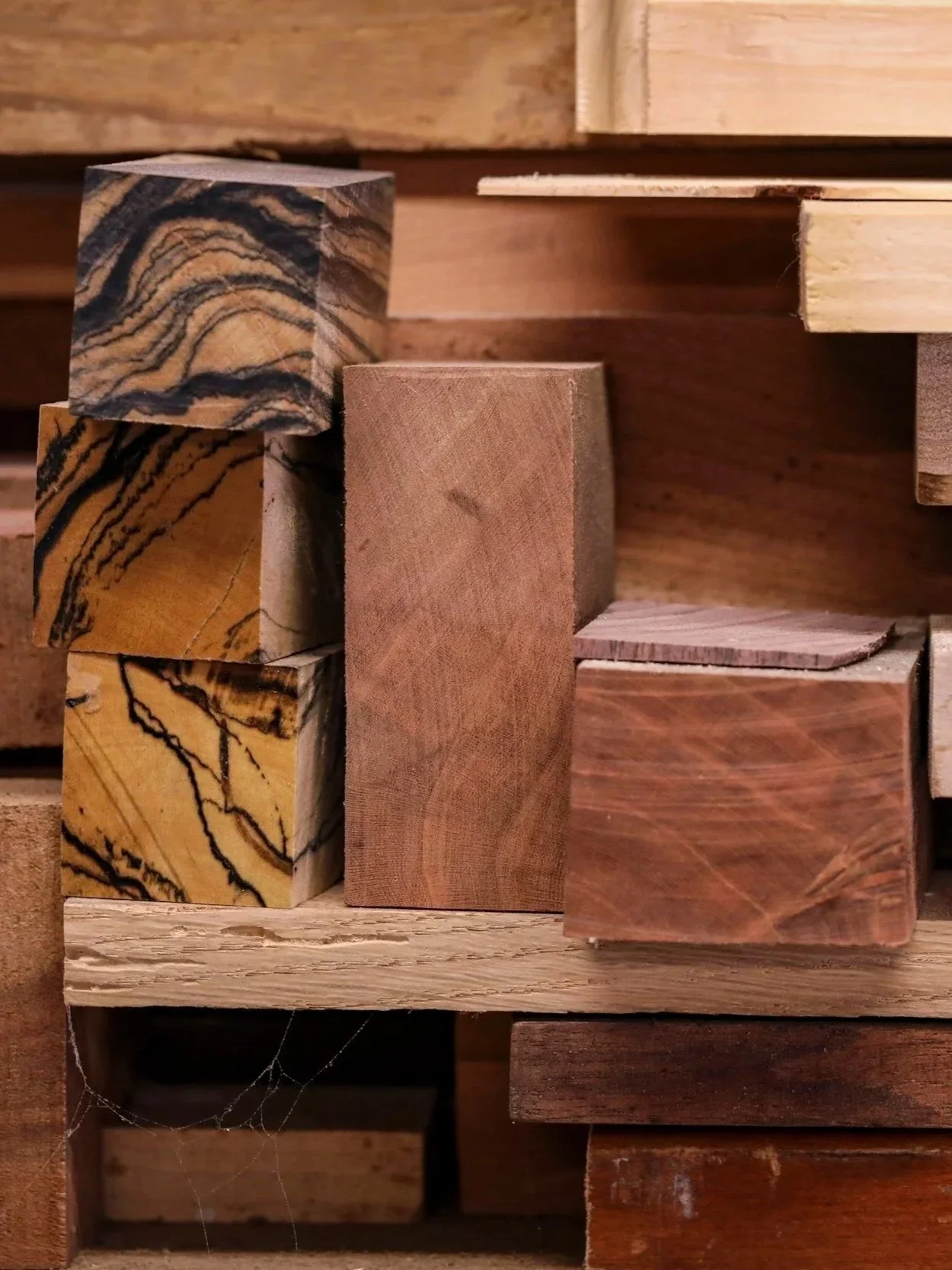 Stacked pieces of various types of wood with different grain patterns and colors, some with visible spider webs in the bottom left corner.