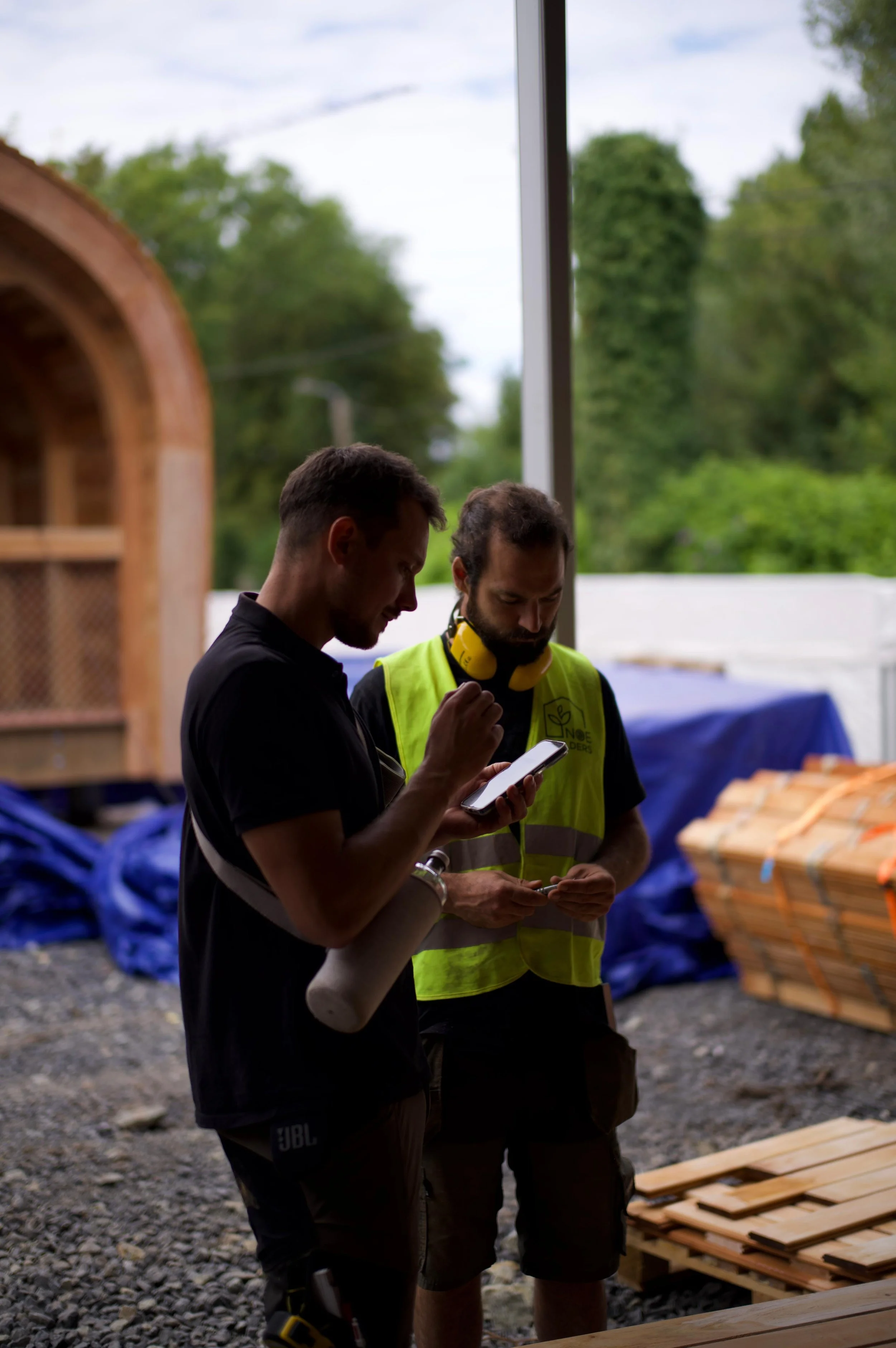 Two men standing outdoors, one in a black shirt and the other in a yellow safety vest with headphones around his neck, looking at their smartphones. Wooden pallets and trees in the background.