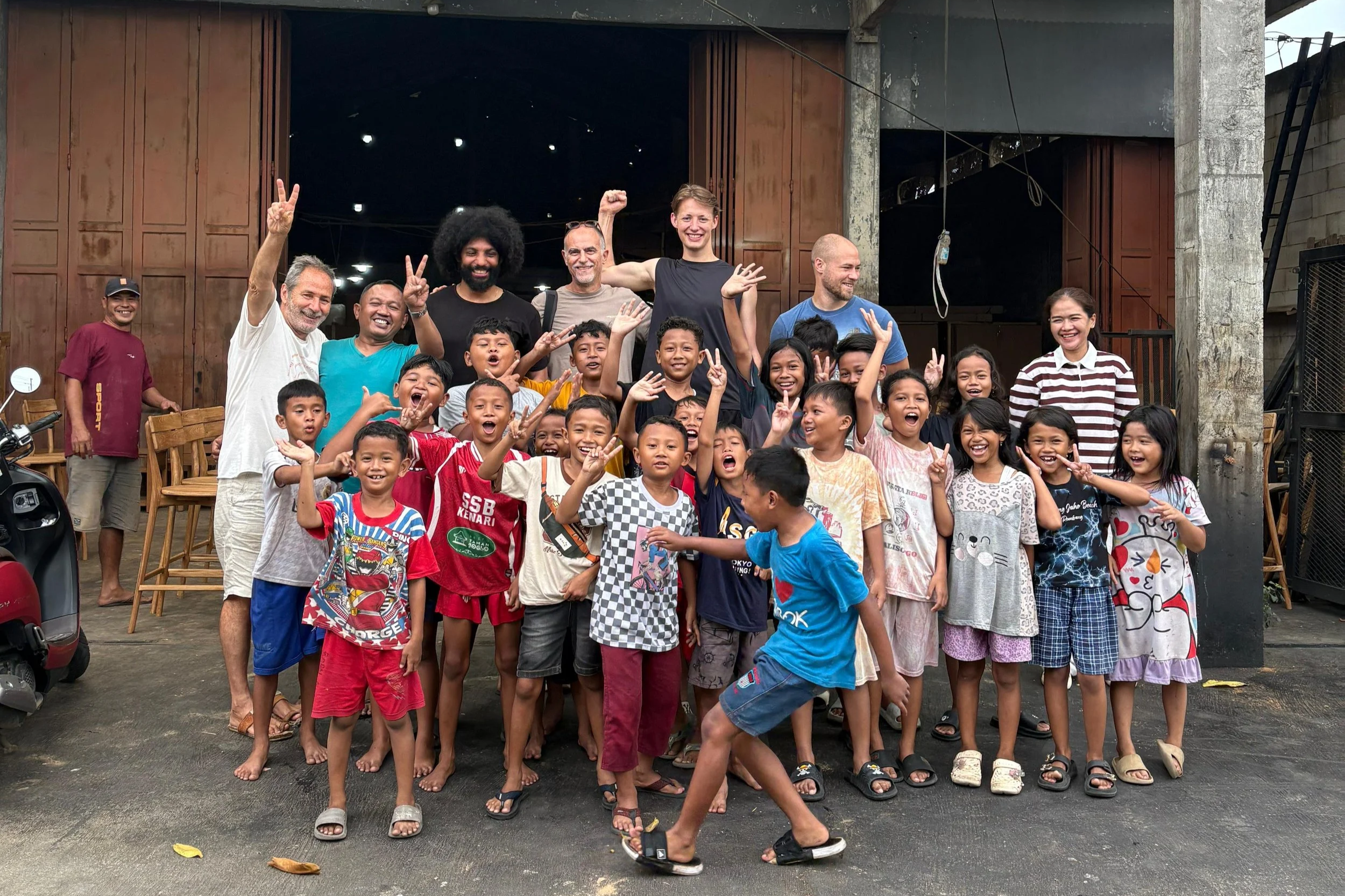 A large group of children and adults smiling and posing for a photo in front of a wooden building or shed. Some children are making peace signs. The setting appears to be outdoors, possibly in a school or community center yard.