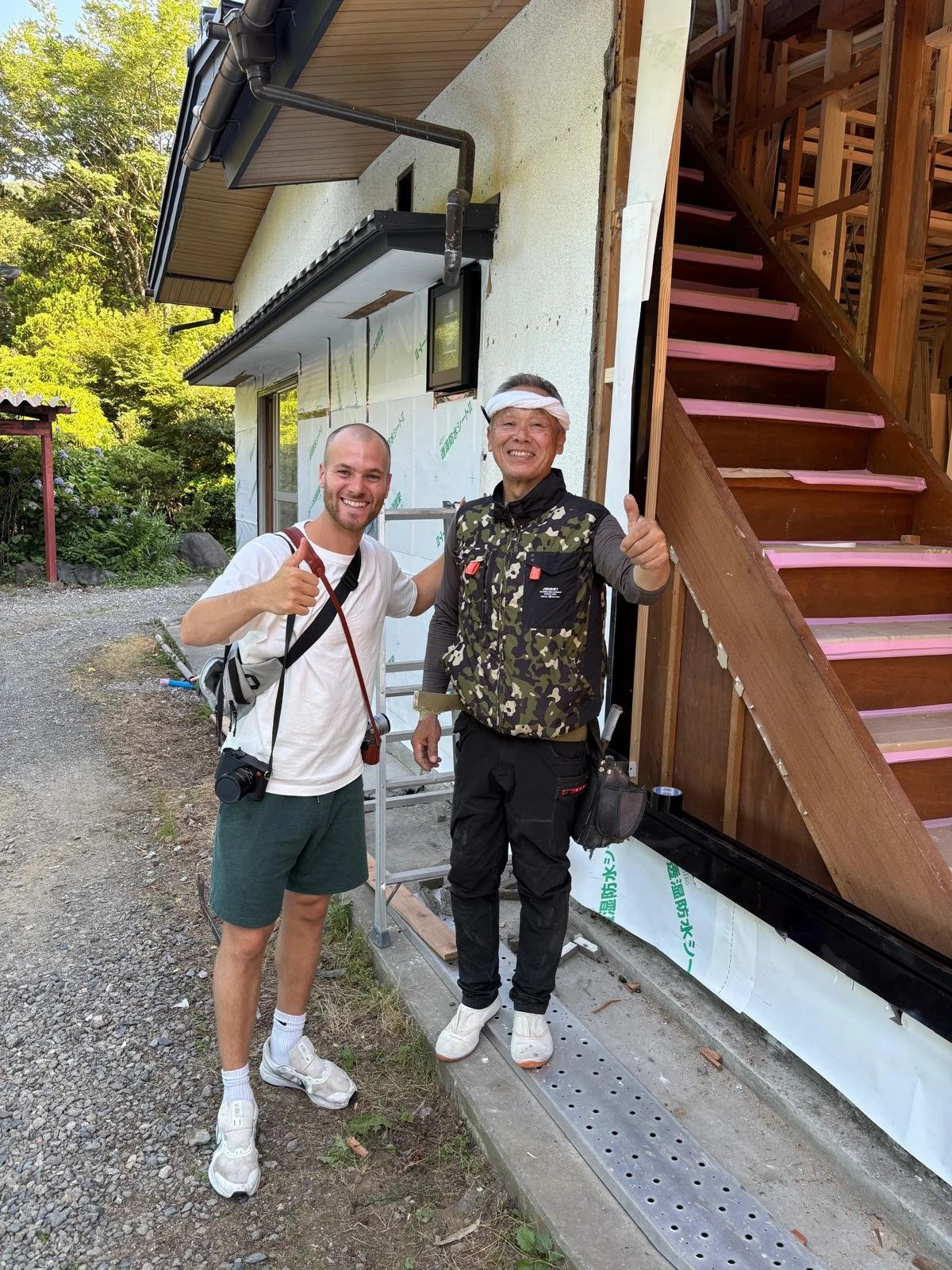 Two men standing outside a house under construction, smiling and giving thumbs up. One is wearing a white t-shirt and shorts with a camera around his neck, the other is wearing a camouflage vest and black pants, with a bandage on his head. The house has exposed wooden stairs and construction materials nearby.