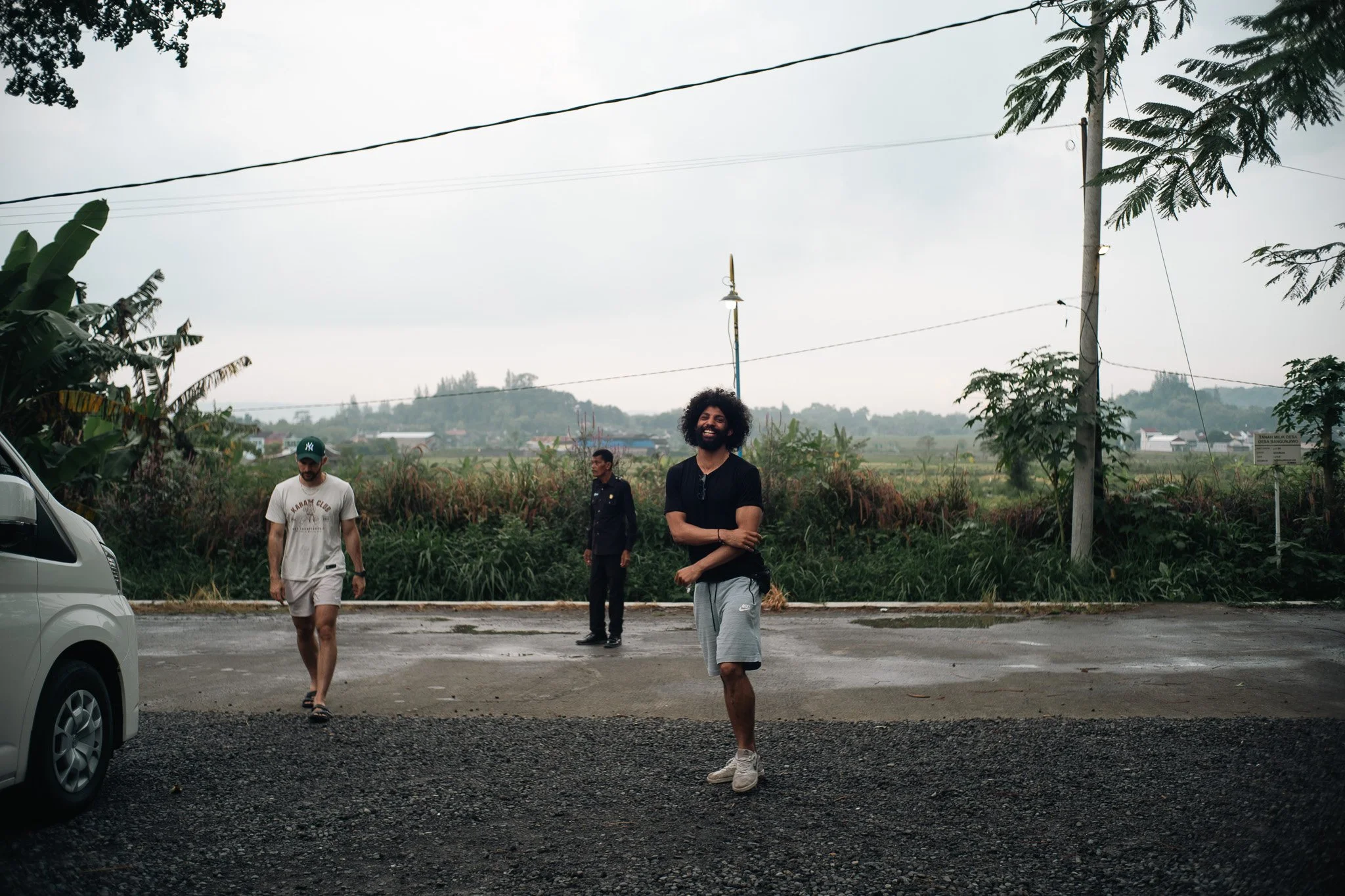 Three men walking on a rural street with a man smiling in the foreground. One man is wearing a black t-shirt and gray shorts, another is wearing a white t-shirt and shorts, and the third man is in a dark shirt. A car is partially visible on the left,