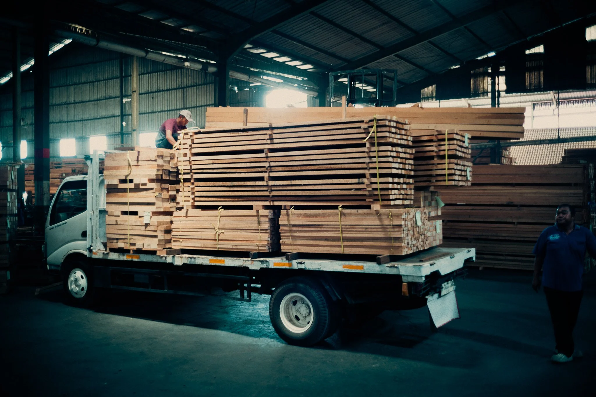 A person loading or unloading a stack of wooden planks onto a flatbed truck inside a warehouse.
