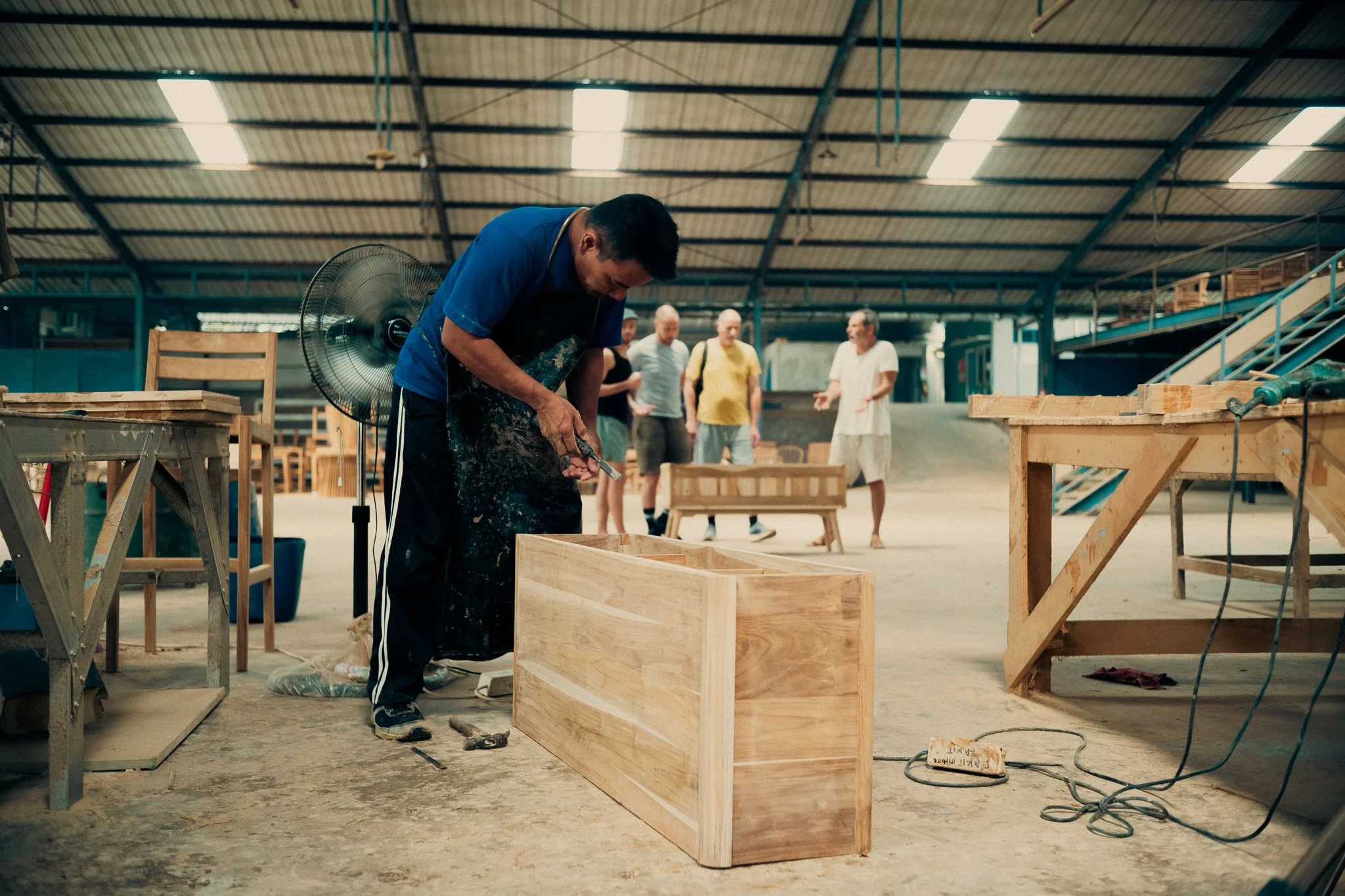 Man working on a wooden box in a woodworking workshop with several people observing.