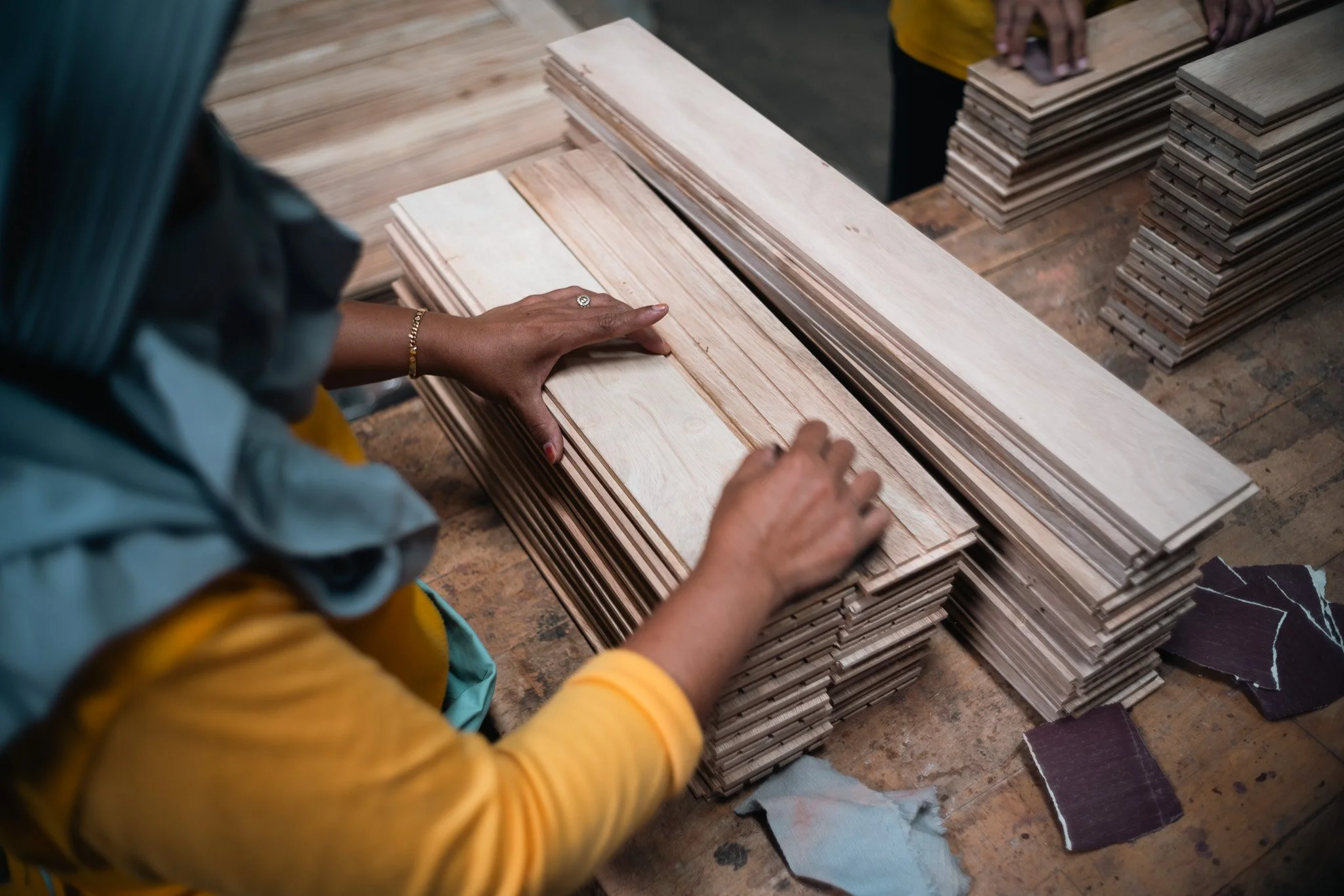 A person examining wooden planks in a woodworking shop, with stacks of wood and sample materials on the workbench.