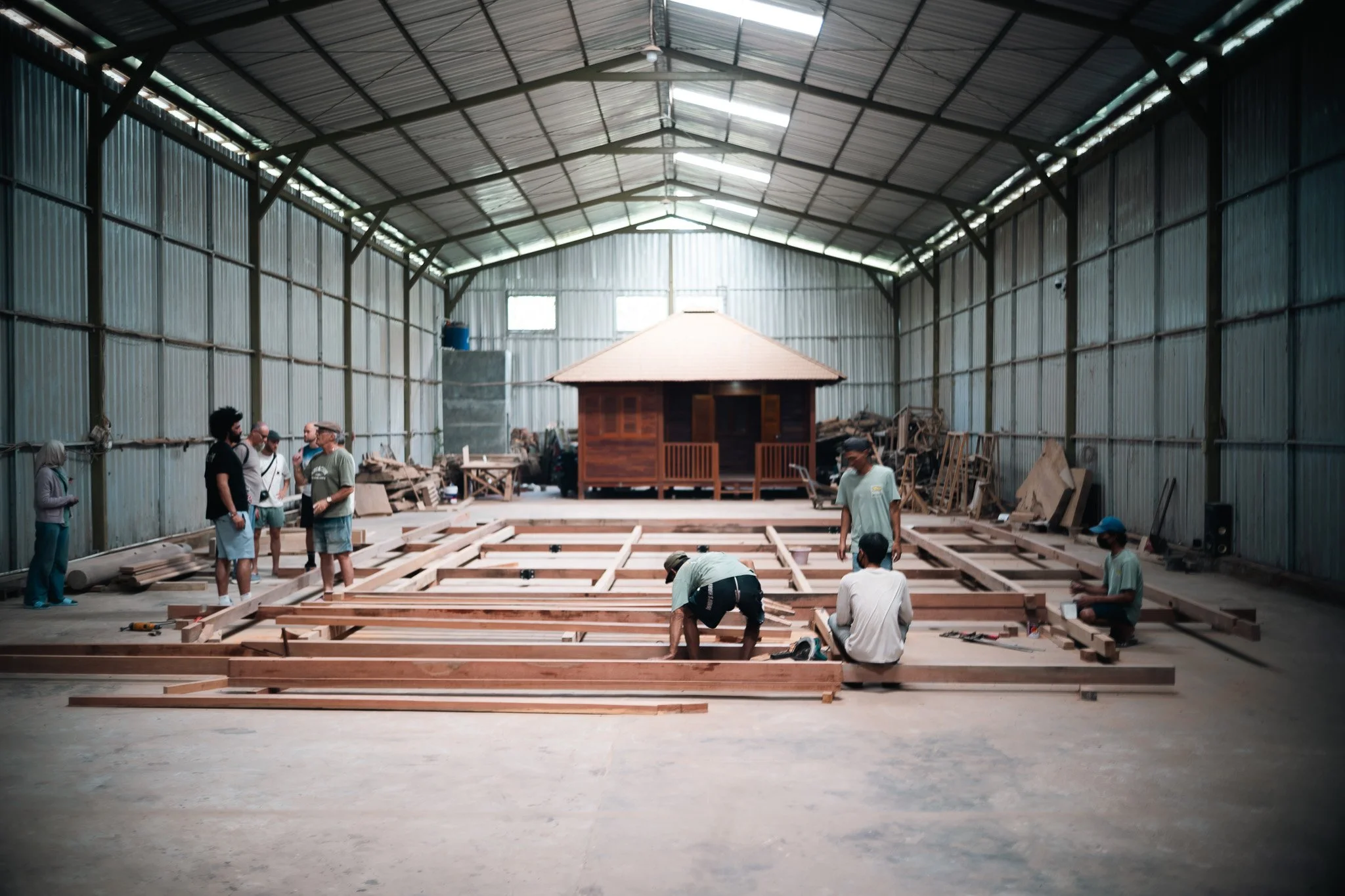 A group of people working together on a construction project inside a large, partially enclosed metal building with a high ceiling. They are assembling a wooden framework for a structure.