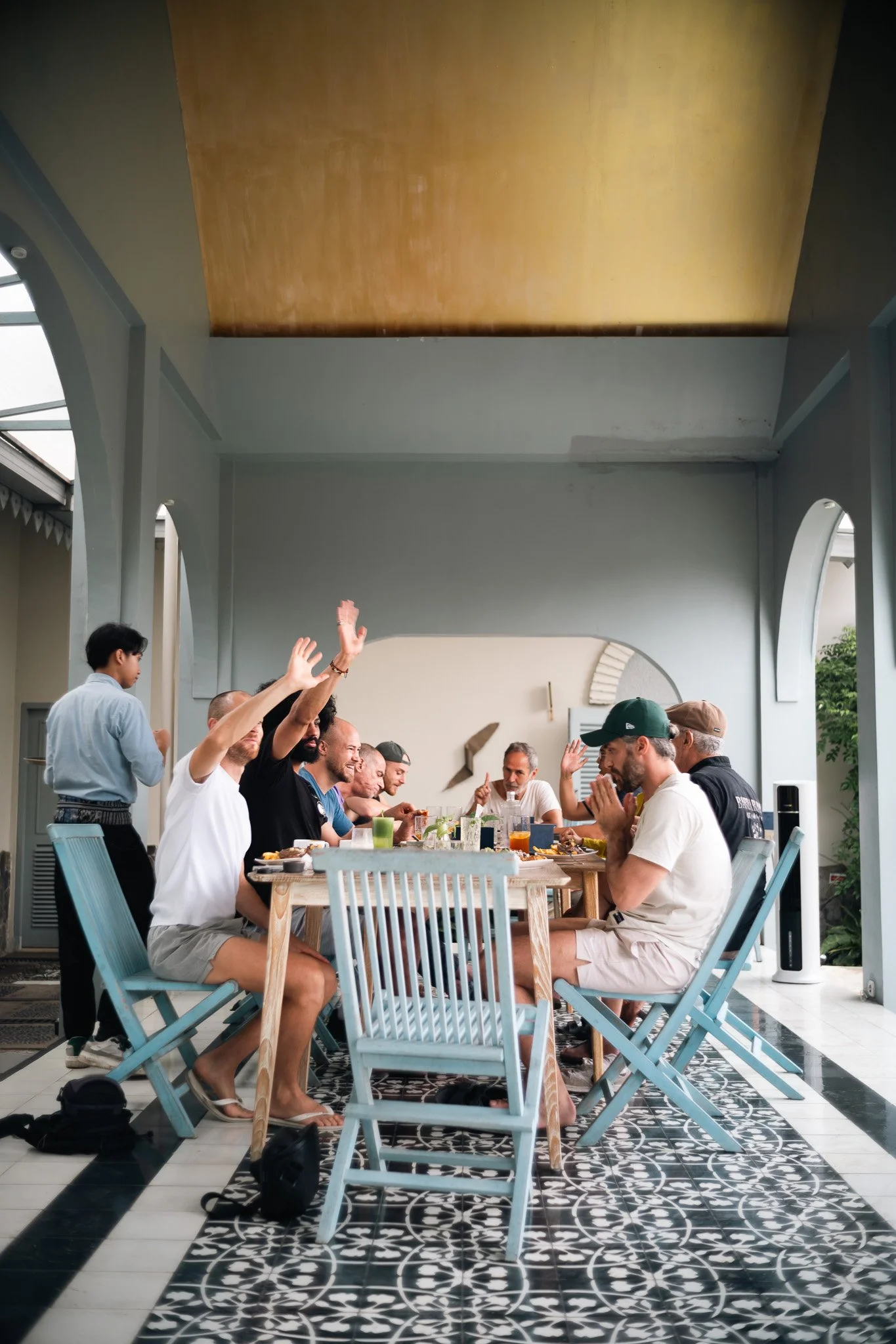 People enjoying a meal at a long table outdoors under a covered patio, some raising hands, with decorative black-and-white patterned floor tiles.