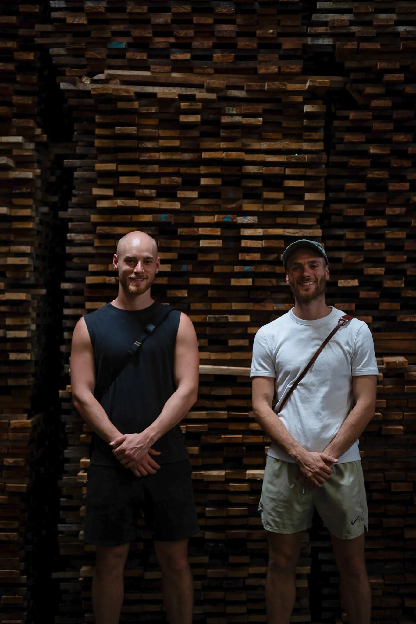 Two men standing in front of a large stack of wooden planks arranged in a pattern and smiling at the camera.