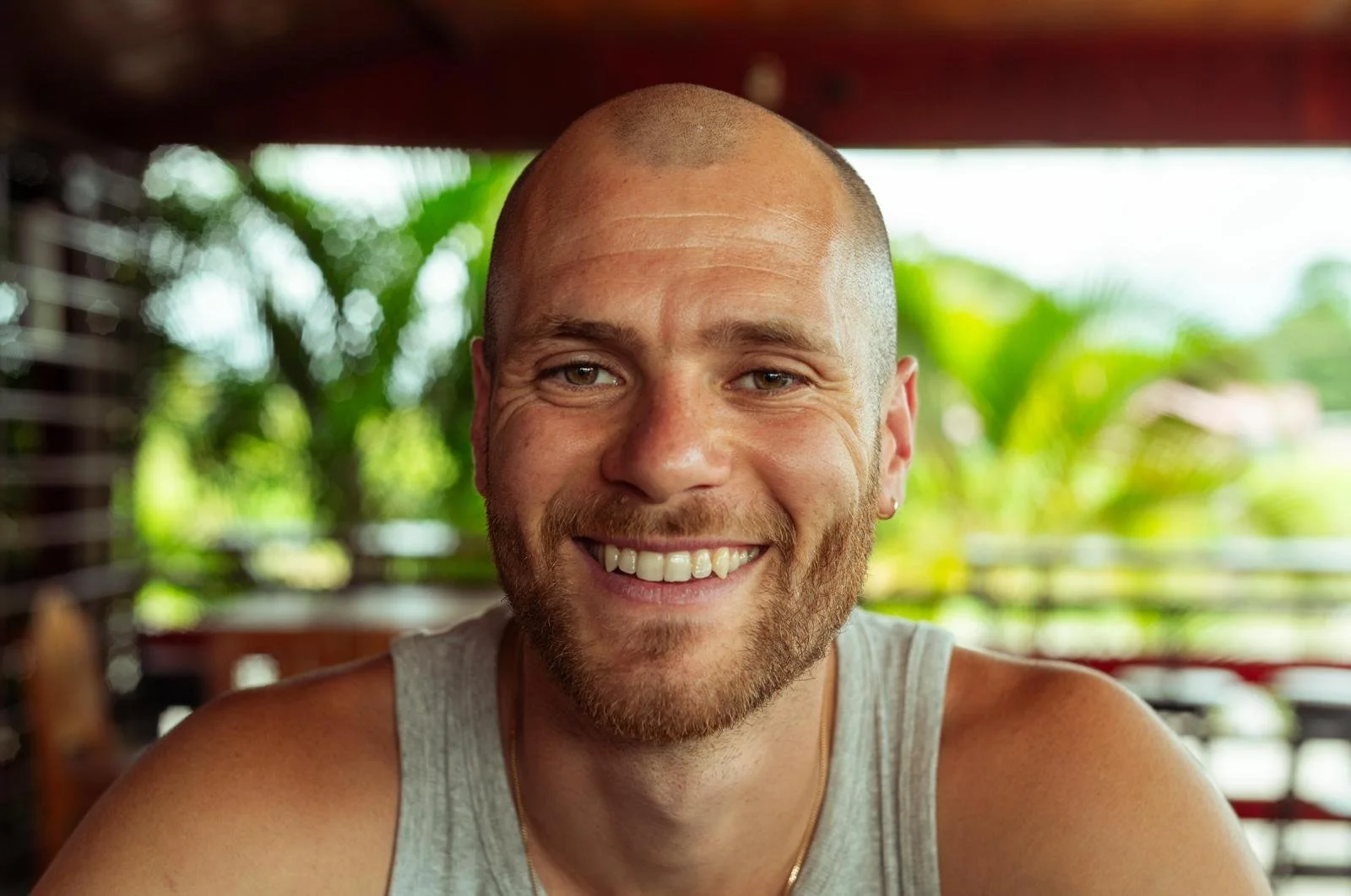 A man with a shaved head, beard, and mustache smiling at the camera. He is wearing a gray tank top and a gold chain. The background is blurred greenery and wooden structures.