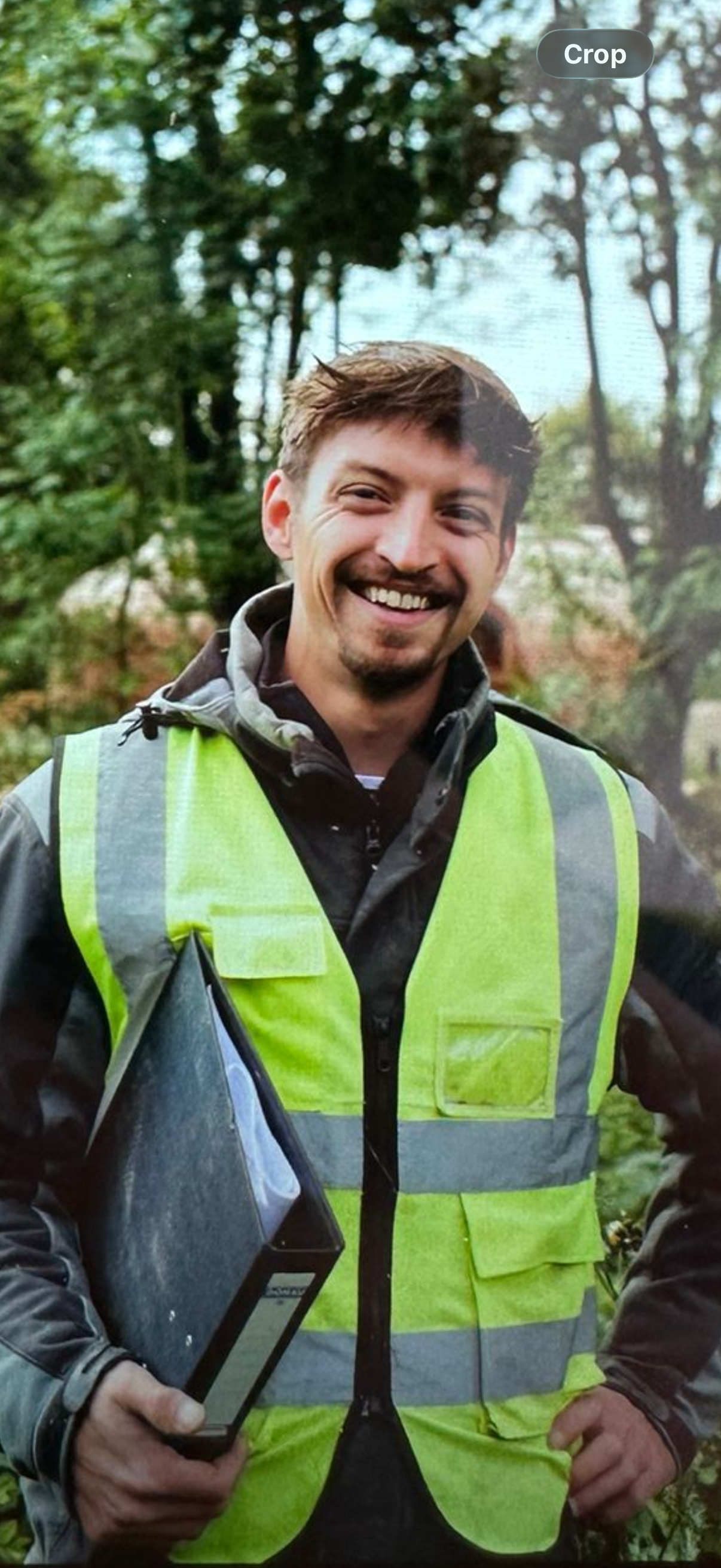 A man wearing a high visibility yellow safety vest and outdoor gear, holding a handheld device, standing in a wooded area outdoors.