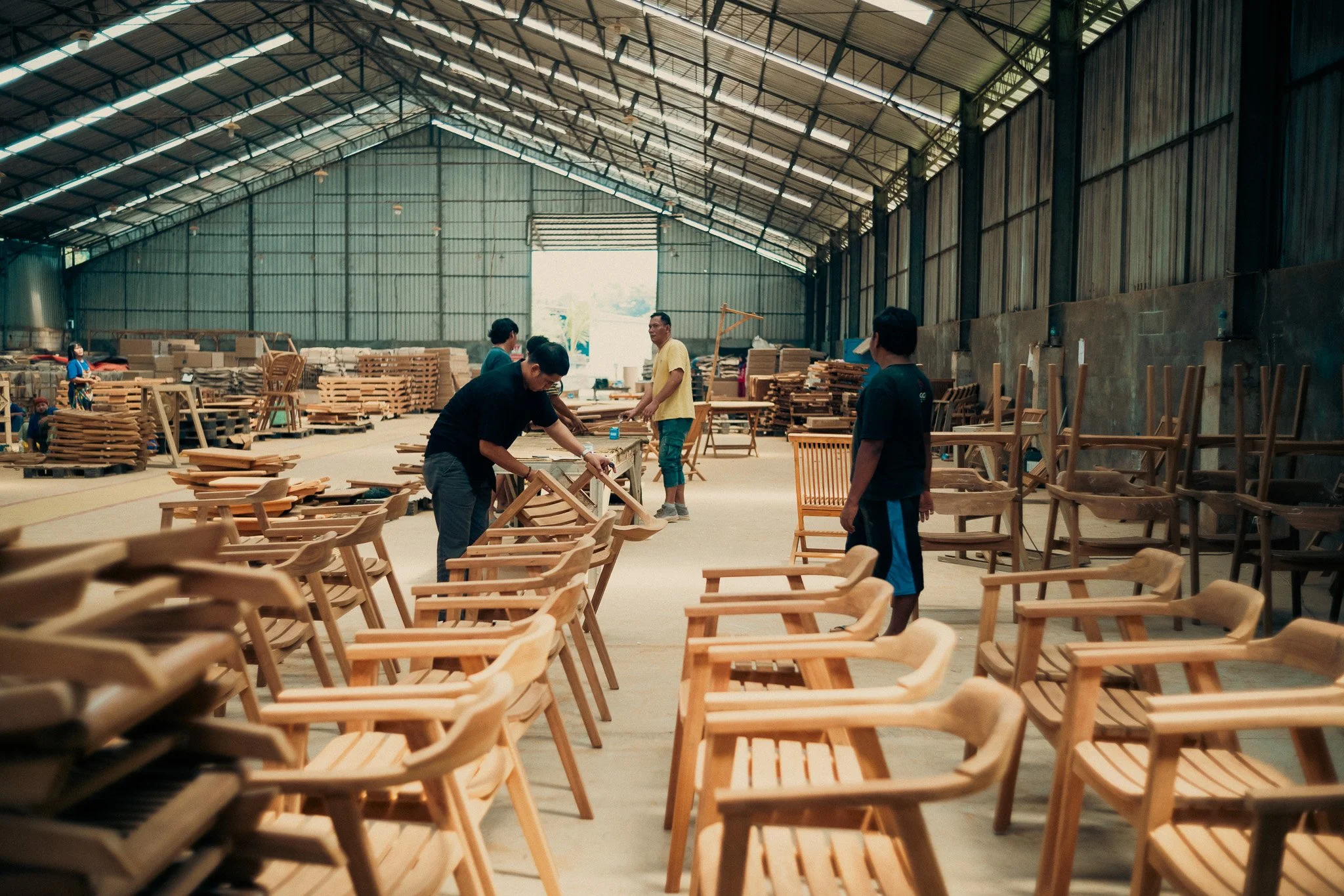 Workers assembling wooden chairs inside a large warehouse with high ceilings and metal walls.