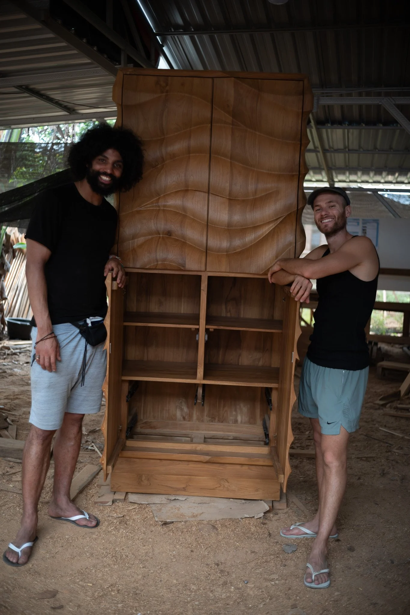 Two men smiling and standing beside a large wooden cabinet in a woodworking shop or workshop. One man has curly hair and beard, wearing a black shirt and shorts, and the other is wearing a cap, tank top, and shorts.