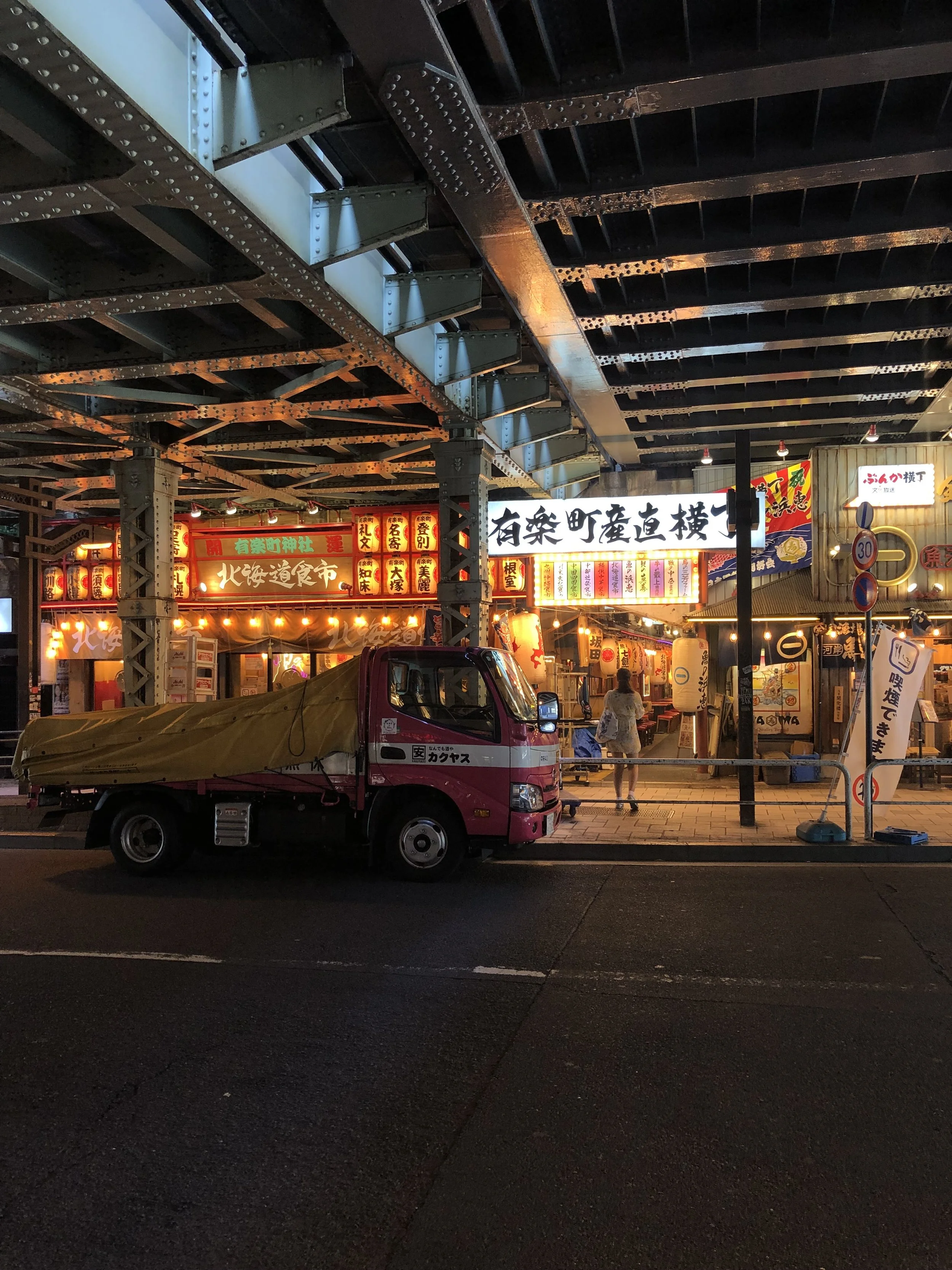 Night scene of a Japanese street with illuminated signs, lanterns, and a pink truck parked by the sidewalk. There are structures overhead, and a woman walking on the street.