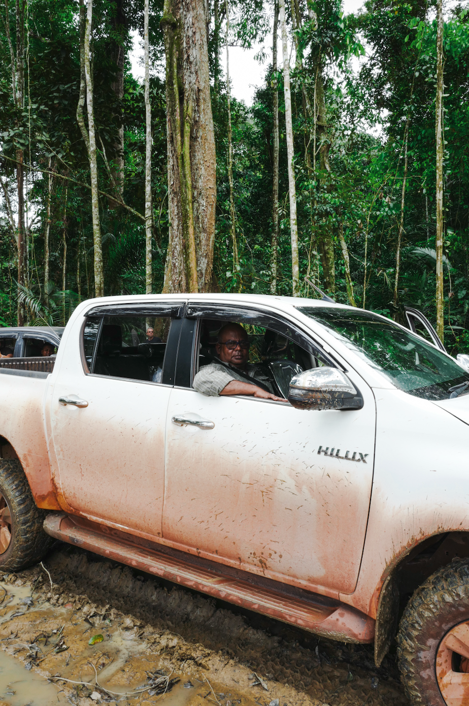 A white Toyota Hilux pickup truck with muddy sides parked on a dirt trail in a dense, green jungle. A man is sitting in the driver's seat, looking out the window. Additional vehicles are visible in the background.