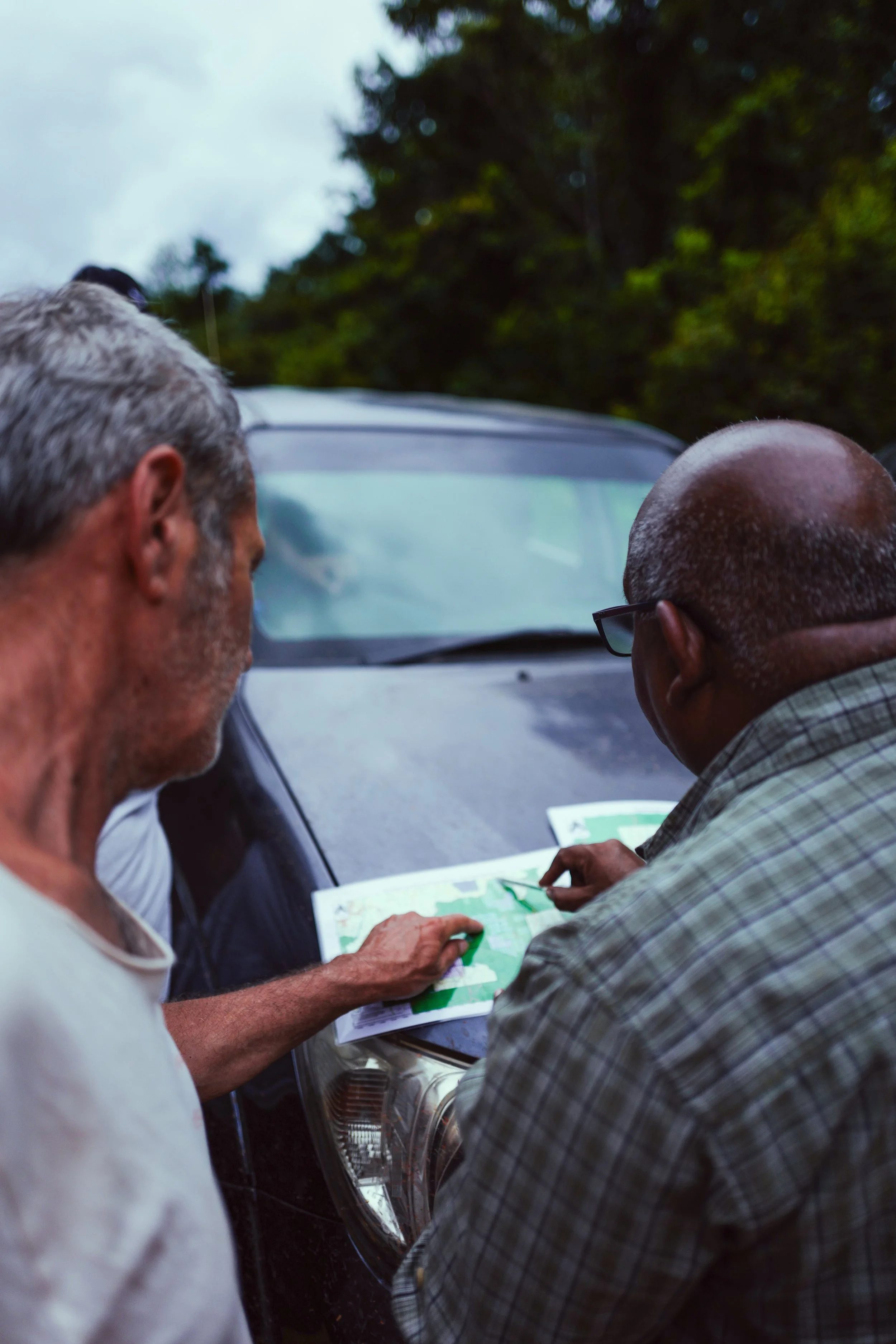 Two men looking at a map in front of a parked car outdoors, with trees and overcast sky in the background.