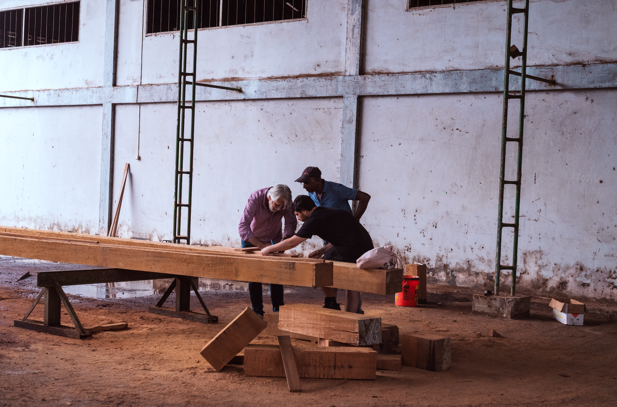 Three people working on a wooden project inside a warehouse or construction site, leaning over a long wooden beam on a workbench, with tools and supplies around them.