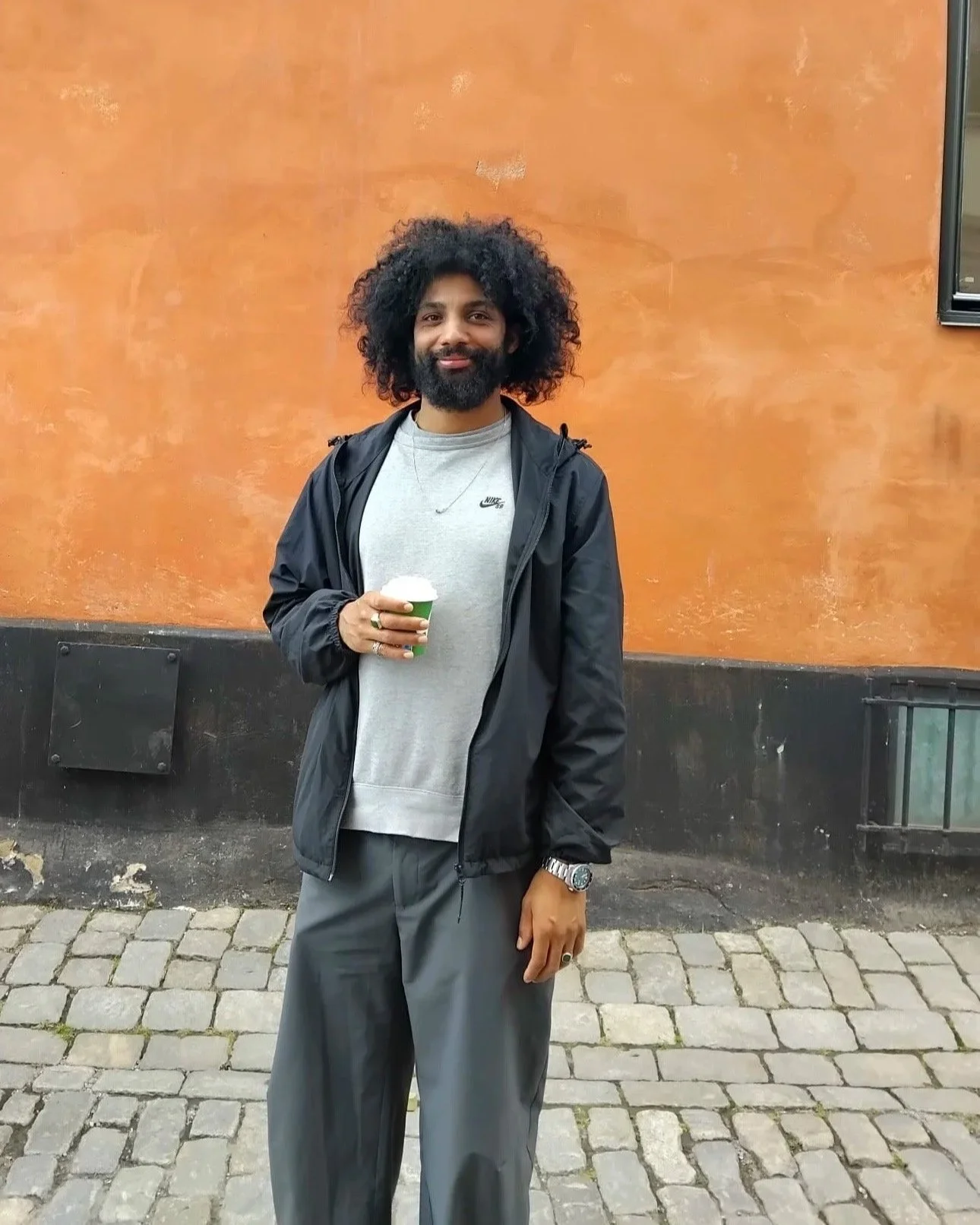 A man with curly dark hair and a beard holding a cup, standing in front of an orange wall on a cobblestone street.