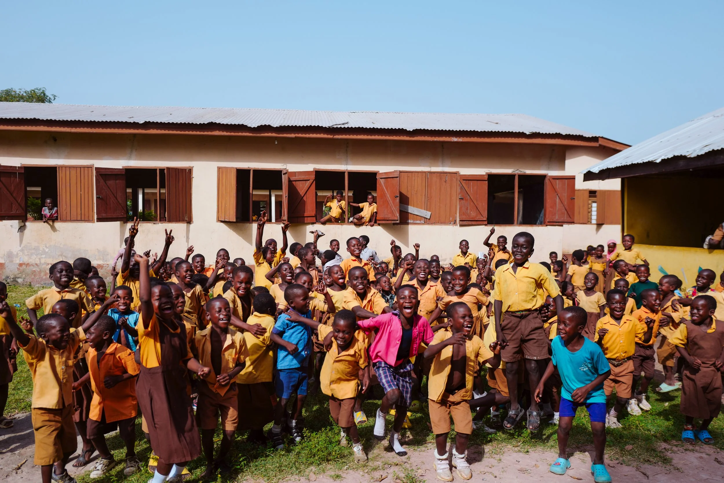 A large group of children in yellow school uniforms smiling, playing, and raising their hands outdoors in front of a school building with open windows and shutters, under a clear blue sky.