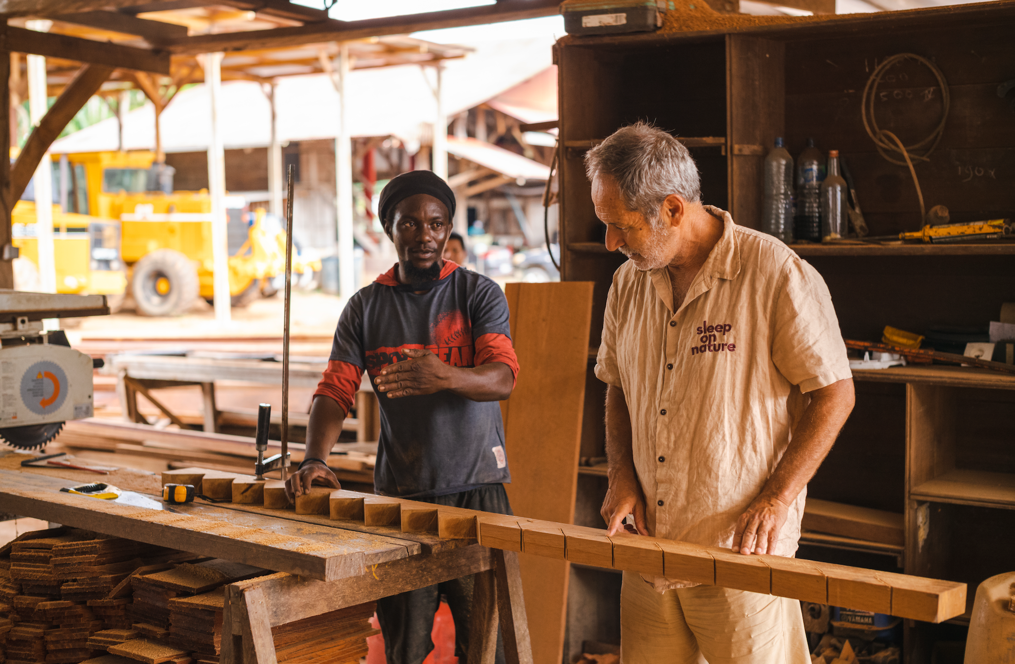 Two men are working together in a woodworking shop. One man appears to be explaining or discussing something, while the other man is focused on a piece of wood. The shop is filled with tools and pieces of wood, with a yellow machinery in the backgrou