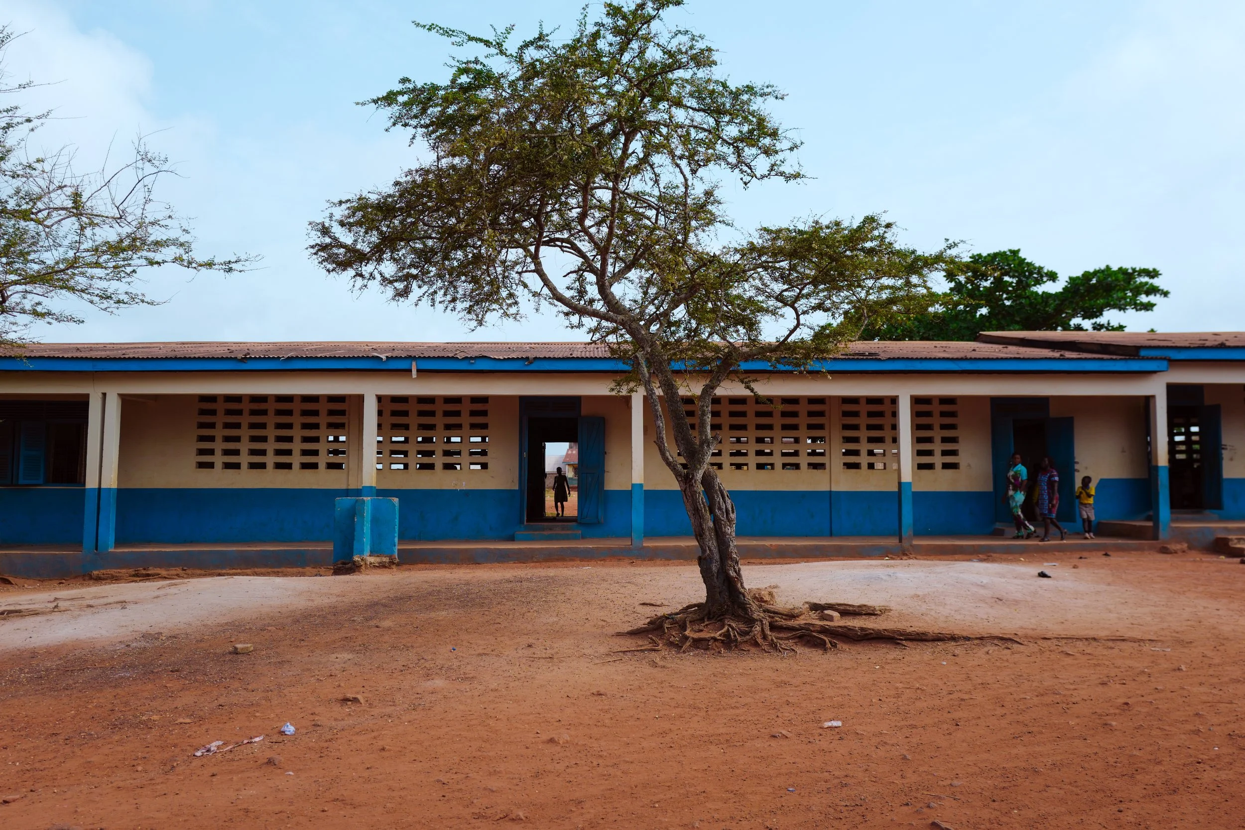 A school building with a large tree in front, people walking outside, and a dirt ground in a rural area.