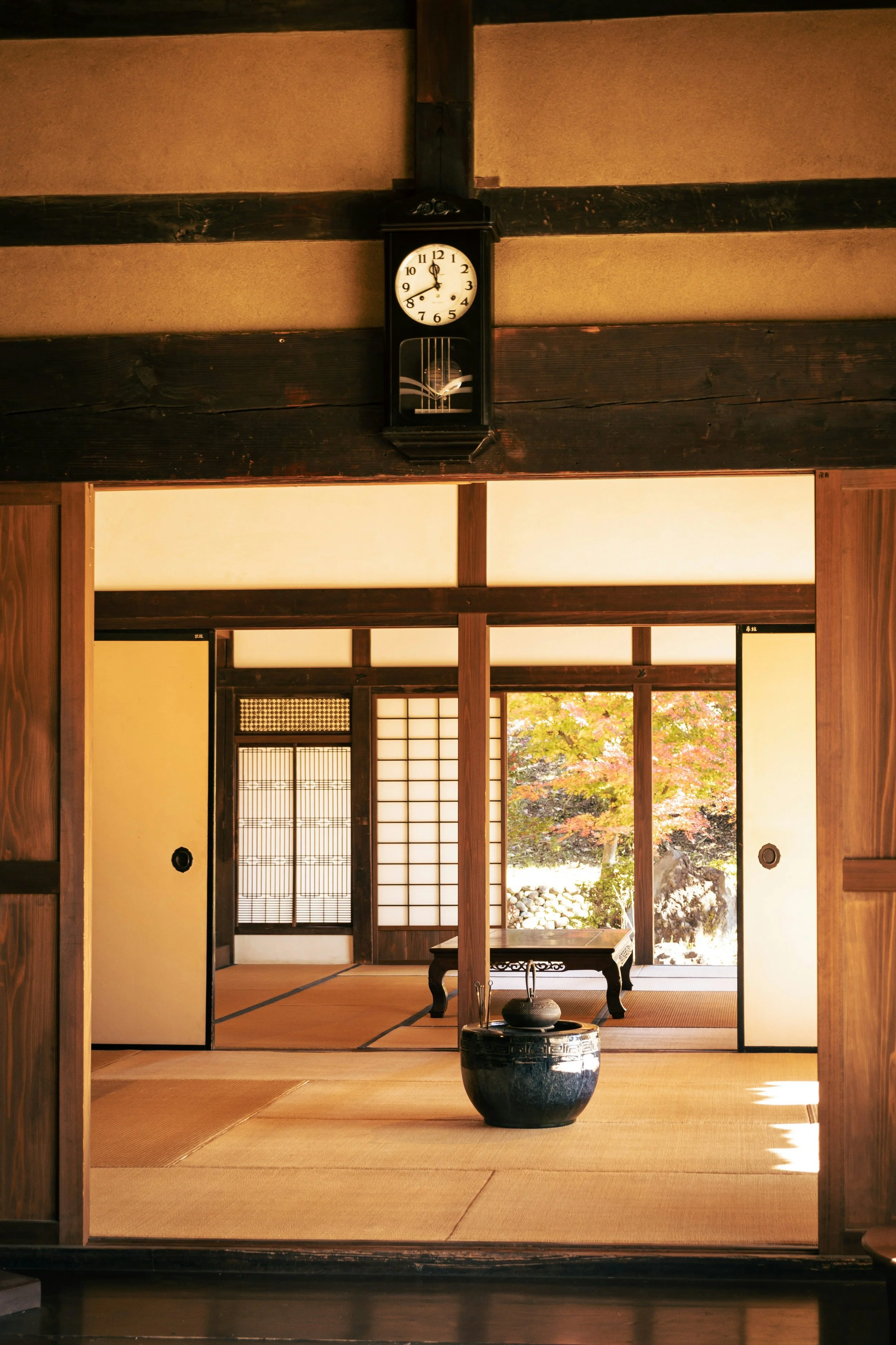 Interior of a traditional Japanese room with tatami mat flooring, sliding shoji doors, and a view of a garden outside. A black wall clock is mounted above the doorway.