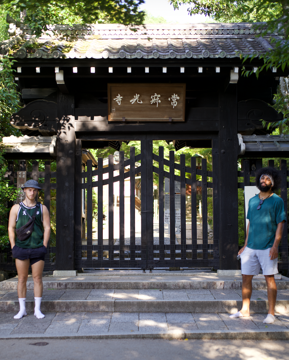 Two men standing in front of a traditional Japanese wooden gate with a wooden sign, surrounded by green trees and shaded sunlight.