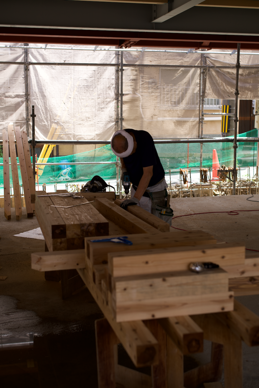 A construction worker with headphones cutting wood on a construction site with scattered lumber and scaffolding in the background.