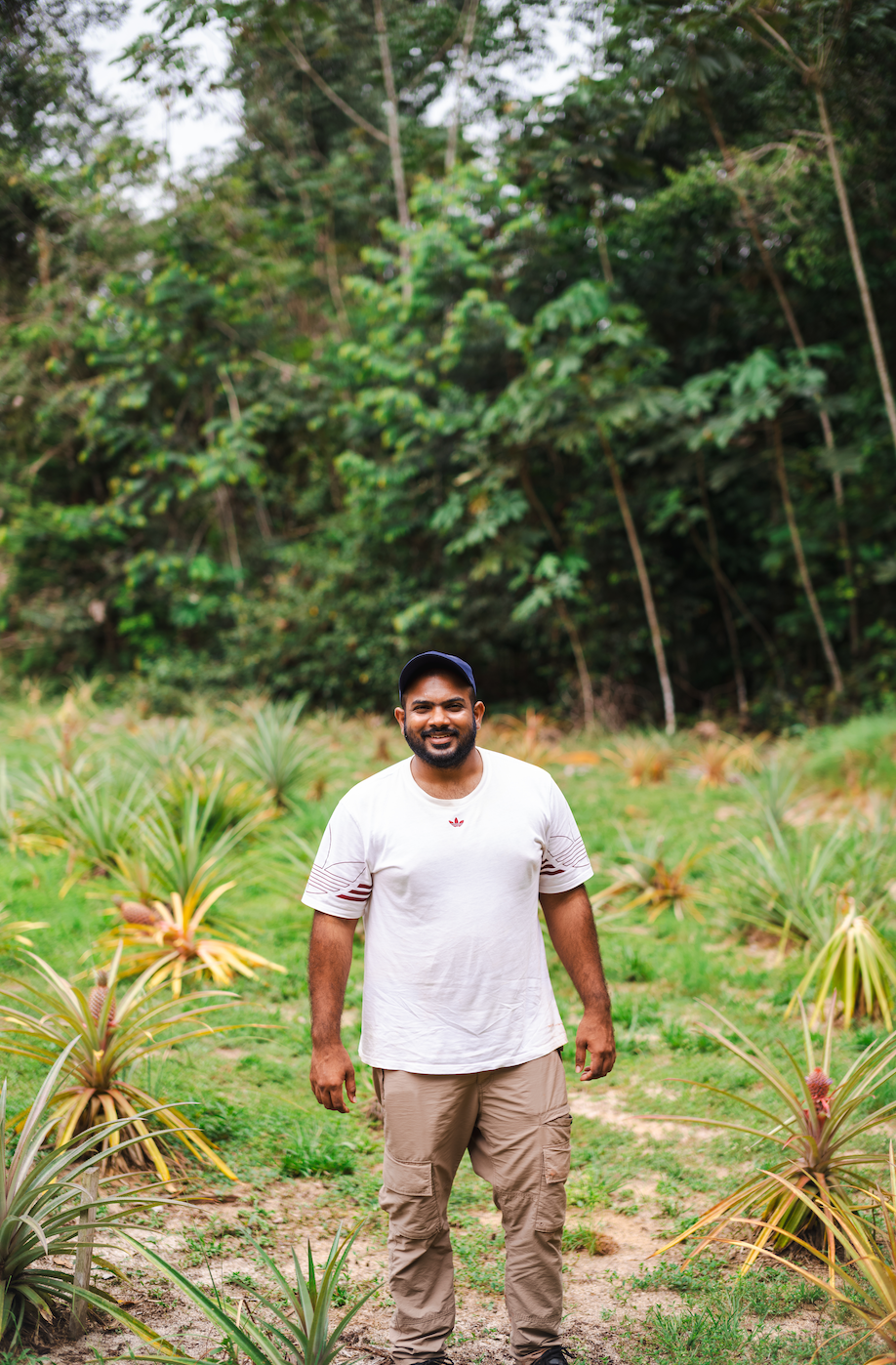 A man stands outdoors in a garden with green plants and trees in the background, smiling at the camera.