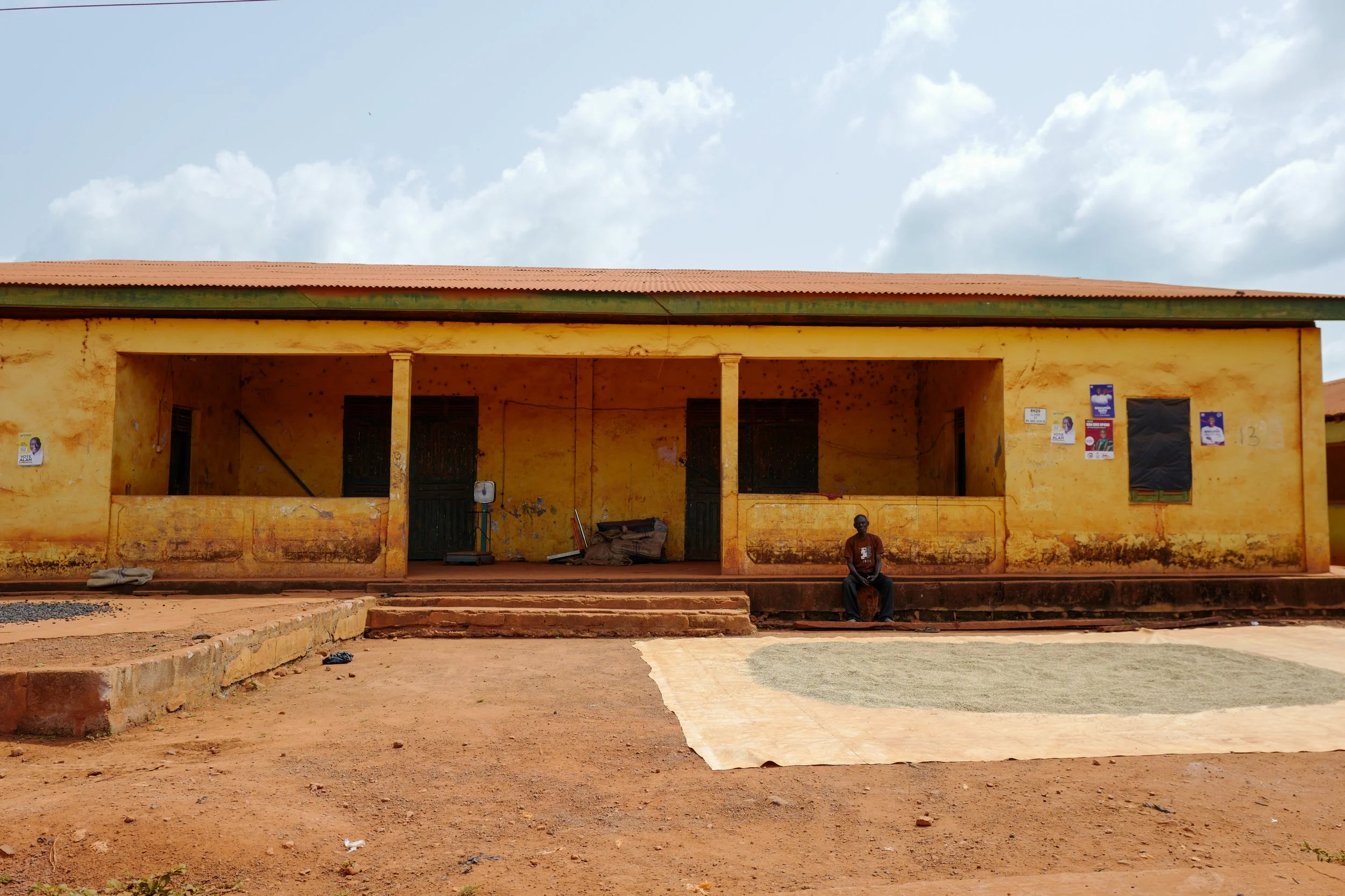 A yellow building with a red-tiled roof and open porch stands against a blue sky with some clouds. There are posters on the right side of the building. A person is sitting on the steps in front of the building.