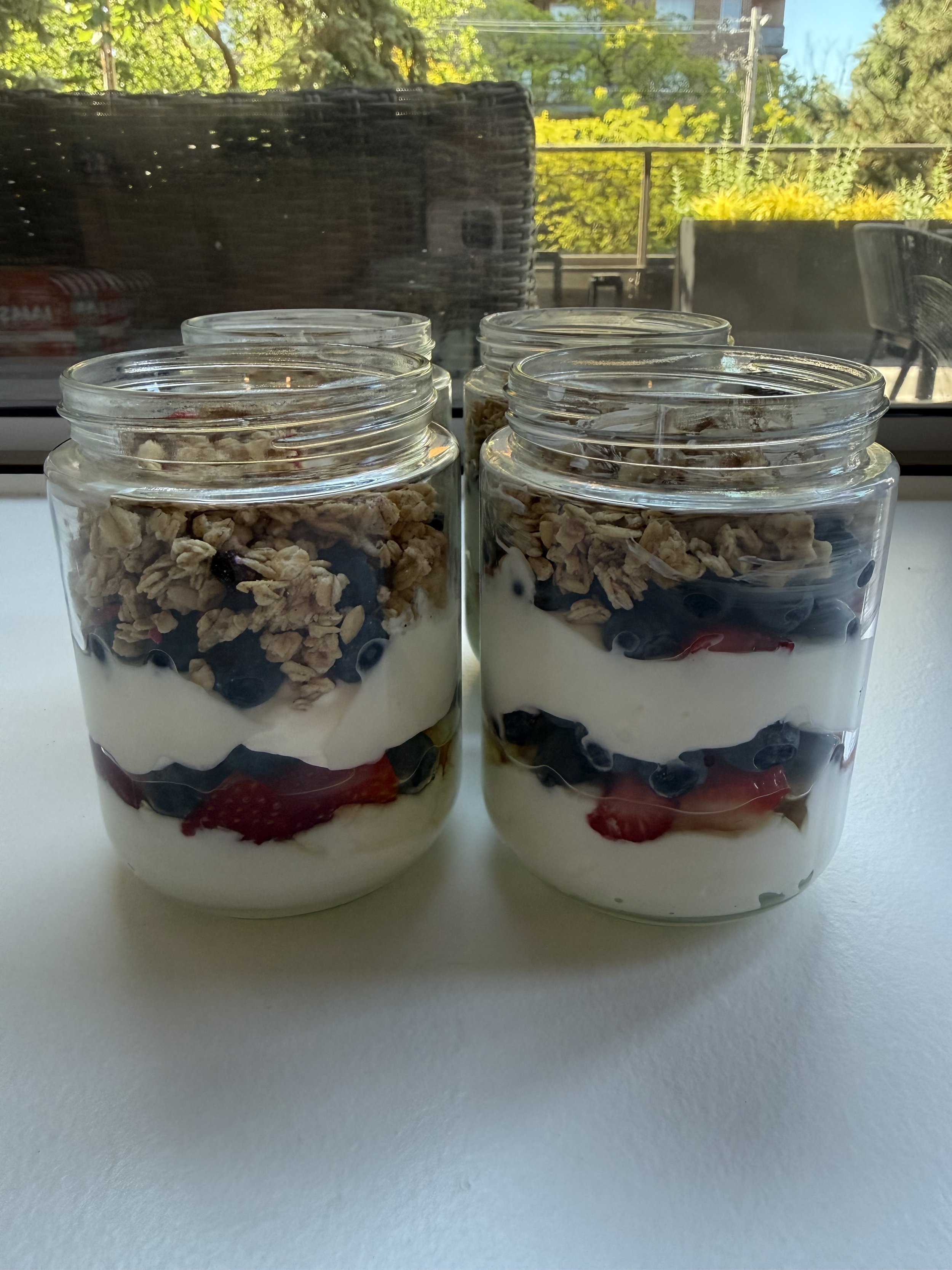 Four glass jars filled with layers of yogurt, granola, blueberries, and sliced strawberries on a white surface with a window showing trees and outdoor seating in the background.
